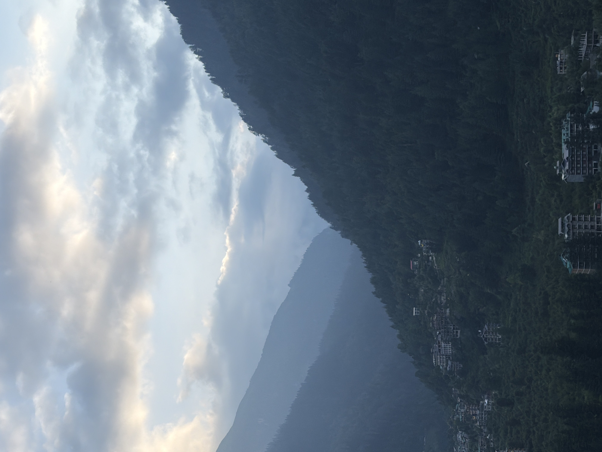 This image captures a panoramic view of the mountainous terrain surrounding Vashisht, India, at dusk. The sky is filled with soft, diffused light from the setting sun, casting a hazy, muted glow through scattered clouds. The prominent feature of the landscape is a series of steep, densely forested mountains that recede into the distance, creating layers of shadow and depth.
On the slopes of these mountains, particularly in the middle ground, small clusters of buildings are visible, indicative of a settlement. These structures appear to be a mix of hotels and residential buildings, built into the hillside amidst the trees. Some of these buildings are multistory and show architectural features common to tourist accommodations. The vegetation is predominantly evergreen trees, forming a dark, textured carpet across the mountainous landscape.
In the foreground, more buildings are scattered across the lower slopes, some with visible roofs and balconies. The overall impression is one of a tranquil, remote village nestled within a vast natural landscape, likely experiencing the cooling and atmospheric conditions of early evening. There are no people or animals clearly discernible in the image.