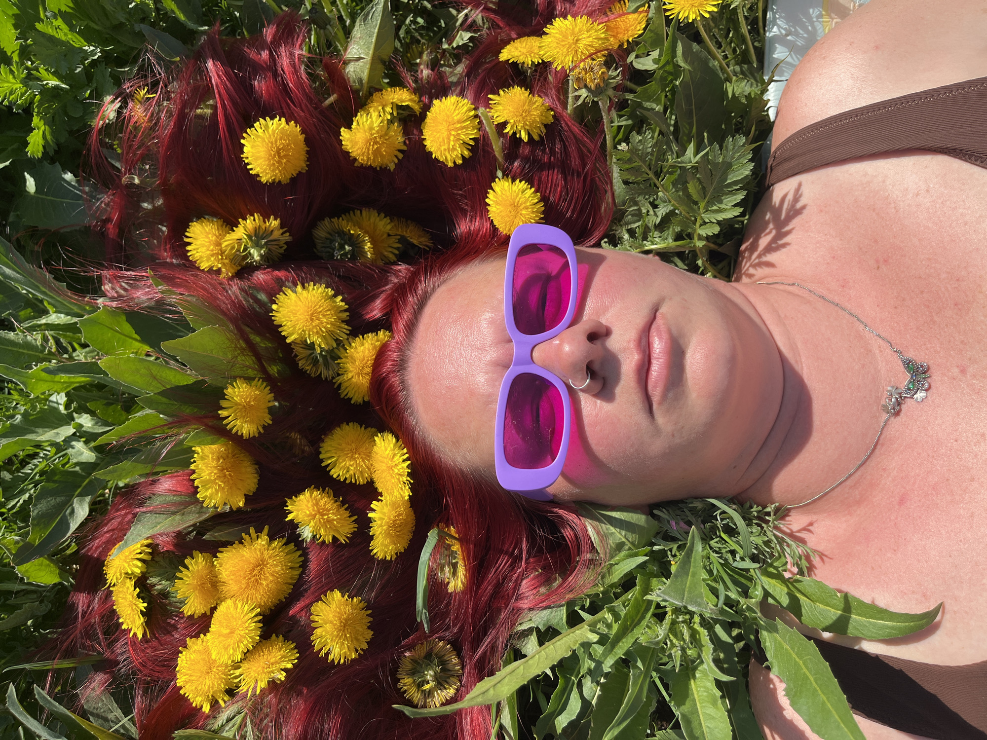 A fair-skinned person with red hair is lying in a field of dandelions in Chaikovsky, Russia, on a sunny day. The hair is adorned with yellow dandelion flowers. The person is wearing lavender-framed sunglasses with dark lenses and a brown top. A silver necklace with a butterfly pendant is visible around their neck. A small hoop is in their nose. The surrounding area consists of dandelions and green plants.