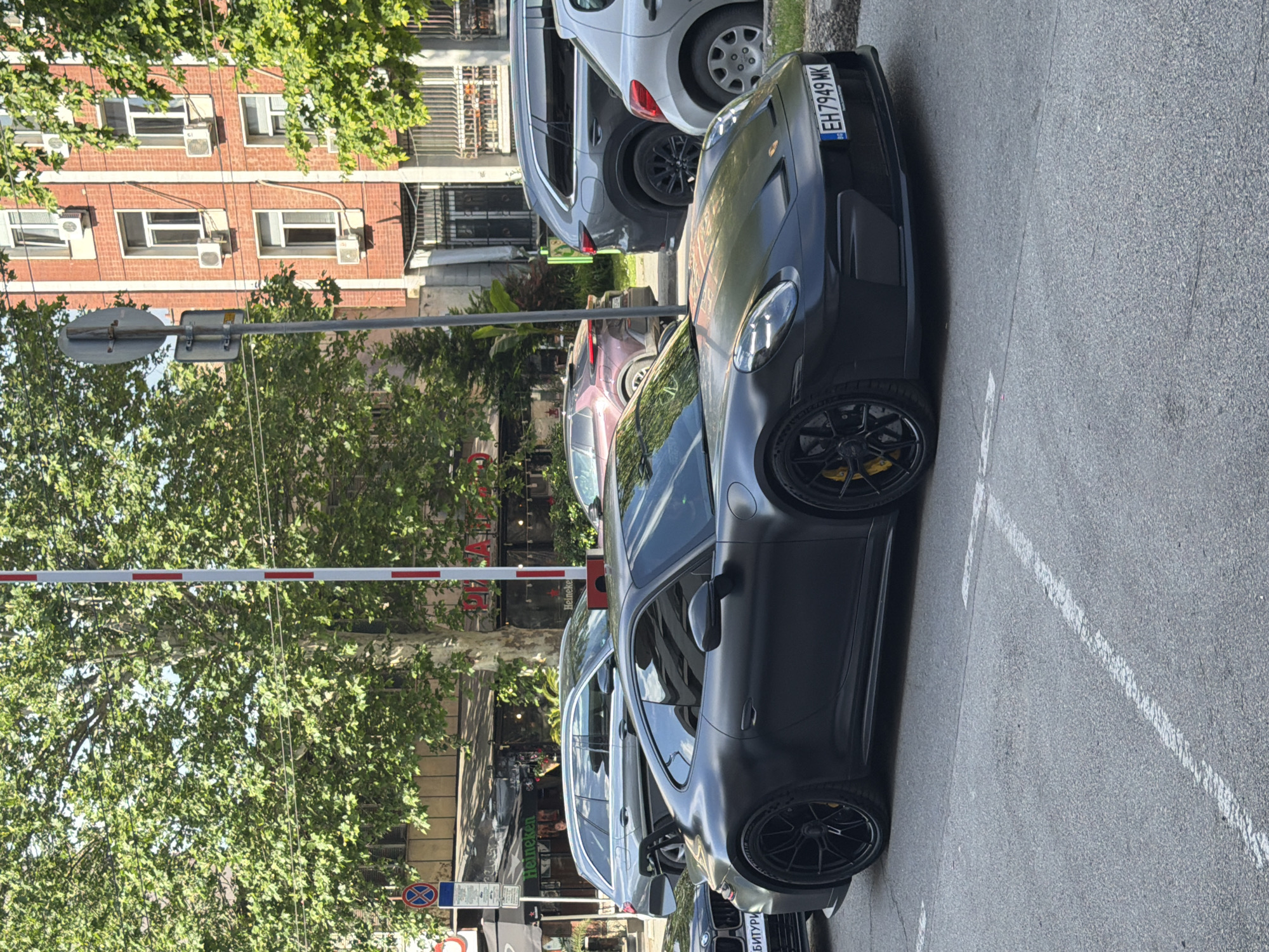 The image depicts a street scene in Pleven, Bulgaria. A matte black Porsche 911 GT3 RS with a Bulgarian license plate (EH7949MK) is parked along the right side of the road. Other vehicles, including a white hatchback, are also parked nearby. The street is lined with buildings, some of which are brick apartment buildings, and trees. A red and white striped barrier is visible along the side of the road. Sidewalk and street markings are also present.