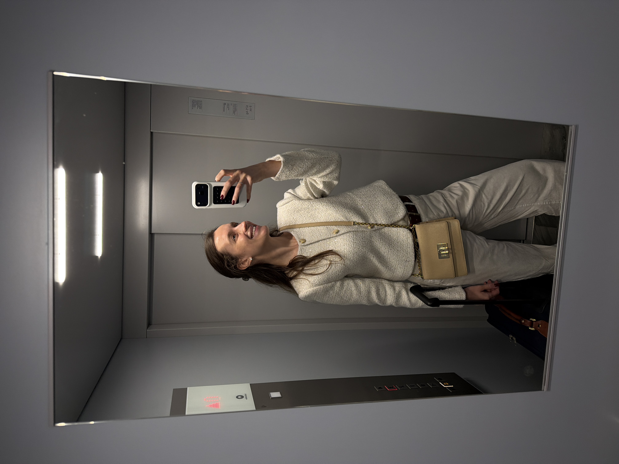 A woman is taking a selfie in a mirror inside an elevator. She is wearing a light-colored tweed jacket, light-colored pants, and a small beige handbag. She is holding a smartphone in her right hand. The elevator interior is gray with a control panel showing floor numbers. A suitcase is visible at the bottom of the mirror. The location is Vozdovac Urban Municipality, Serbia.