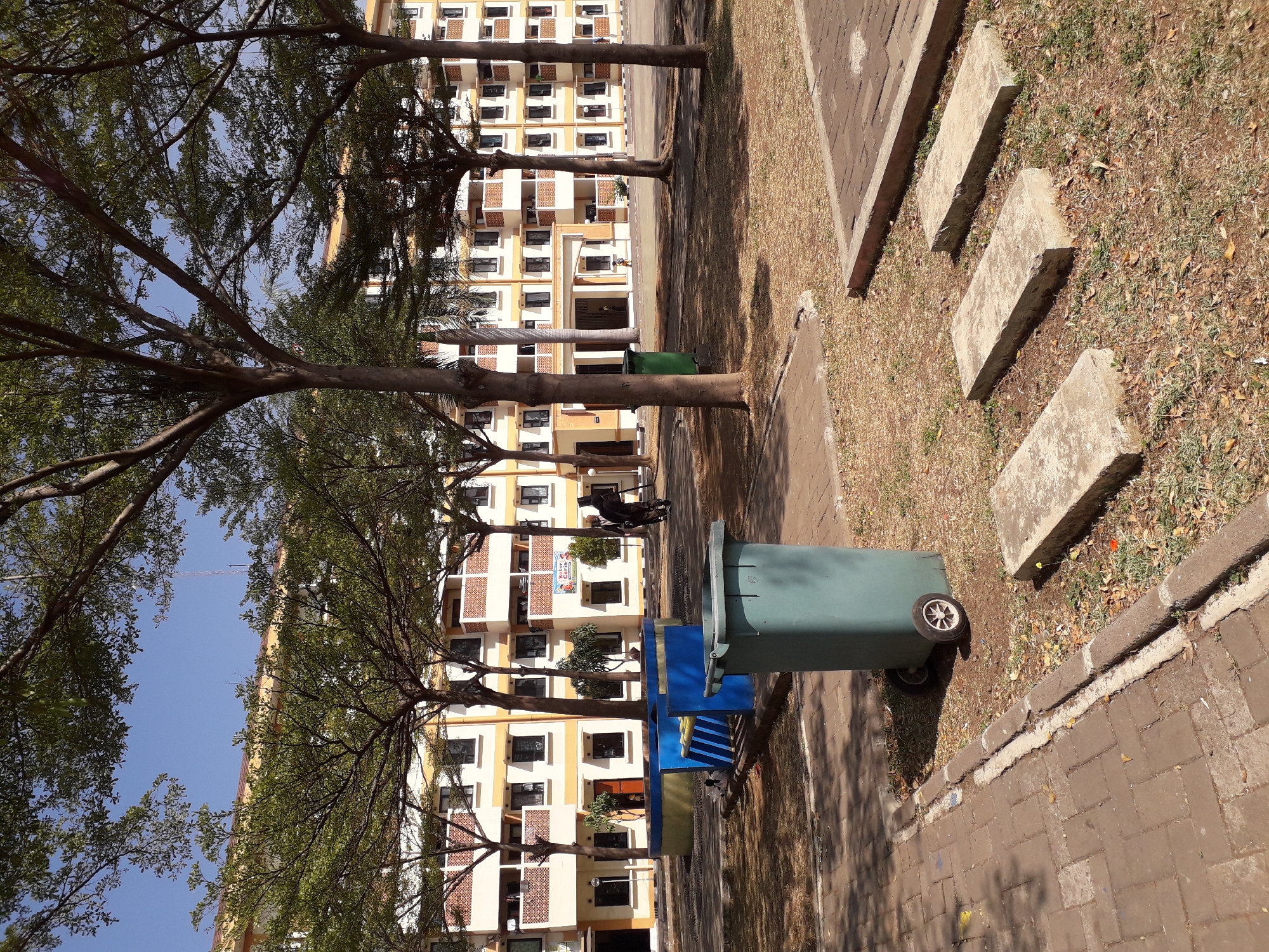 In the city of Cankuang, Indonesia, a residential building with cream and yellow facades stands under a clear blue sky. Several mature trees with sparse foliage are scattered throughout the scene, casting dappled shadows on the ground. In the foreground, a large green trash can with wheels sits on a paved area. To its left, a blue and yellow structure that appears to be a planter or seating area is visible. Behind these, two individuals are walking away from the camera. The ground is covered with dry grass, fallen leaves, and paving stones. The building features multiple levels with balconies and windows. A banner with Indonesian text is displayed on one section of the building.