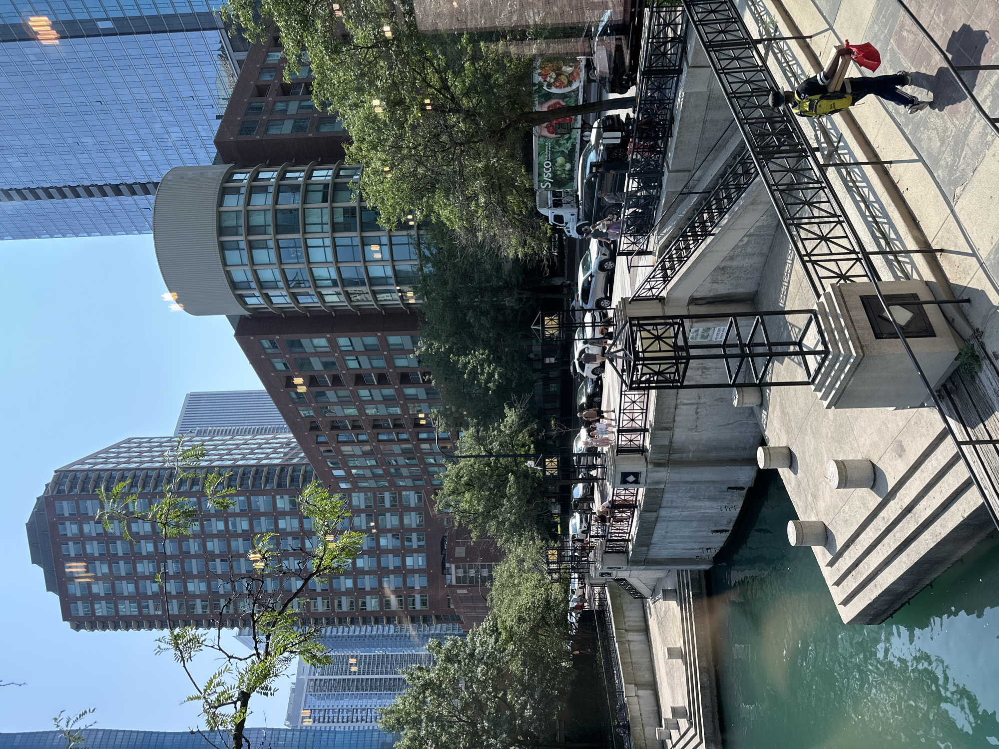 The image depicts a sunny day scene along a canal in Chicago, Illinois. In the foreground, a concrete walkway lines the turquoise canal, with steps leading down to the water's edge. A person wearing a backpack and carrying a red bag and a flat package walks away from the viewer along the elevated walkway. Adjacent to the canal, a stone wall and staircase lead up to a street level.
On the street level, several people are visible walking and standing near parked cars. A white Sysco truck with food imagery on its side is parked among other vehicles, including SUVs and sedans. Beyond the street, a series of multi-story buildings dominate the skyline. These include a tall, brick-clad residential building with numerous windows, a cylindrical building with a light-colored top and curved windows, and a modern glass skyscraper. Lush green trees are interspersed throughout the scene, providing shade and softening the urban landscape. The overall impression is of a vibrant city environment with a prominent waterway and modern architecture.
