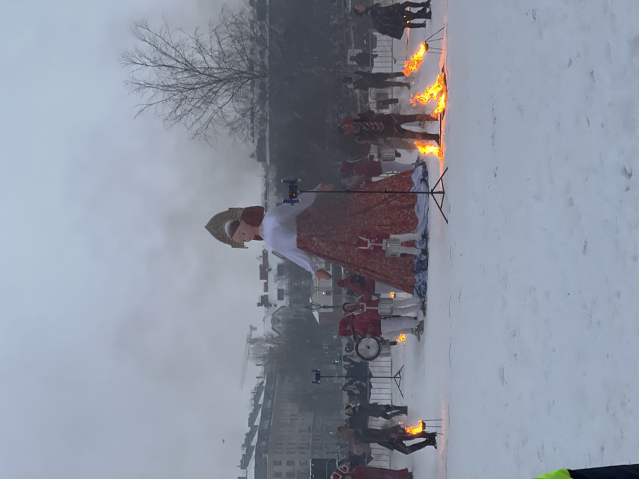 A large effigy of a woman in a white blouse, red patterned skirt, and ornate headwear stands prominently in a snowy outdoor setting. Men in red uniforms stand in the snow around the effigy. Flames rise from torches positioned near the men and puppet. Buildings and bare trees are visible in the background under a cloudy sky. The photograph captures a public display or performance, possibly a historical or cultural event.
