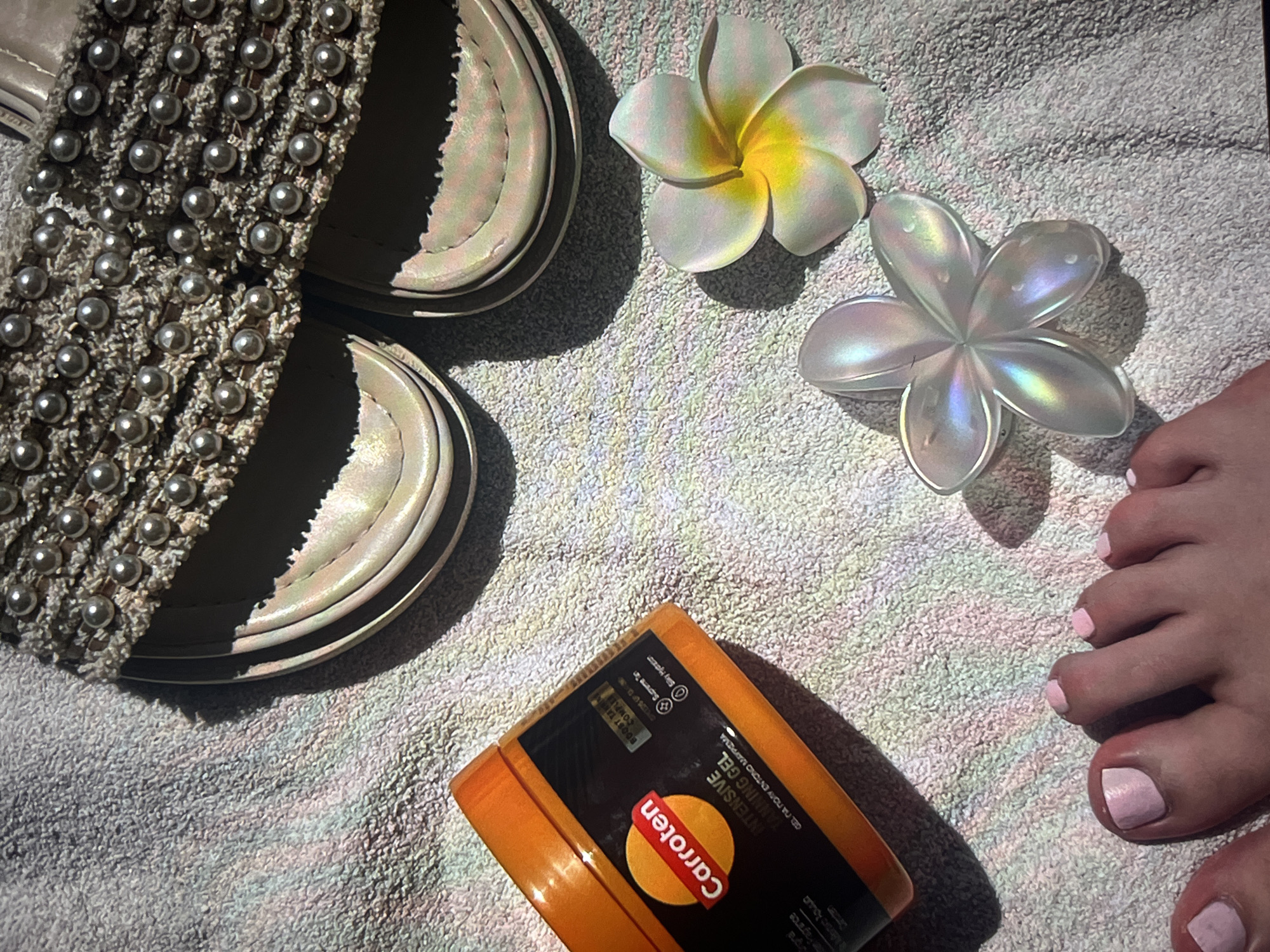 The image shows a close-up shot taken on a textured towel, likely indoors. In the upper left are women's sandals with pearl embellishments. Next to the sandals are two decorative flowers, one a classic plumeria with white petals and a yellow center, and the other a translucent white flower with iridescent sheen. In the bottom left corner is an orange container of "Carroten" intensive tanning gel. To the right side is a person's foot with pink nail polish on the toes. The foot is partially visible.