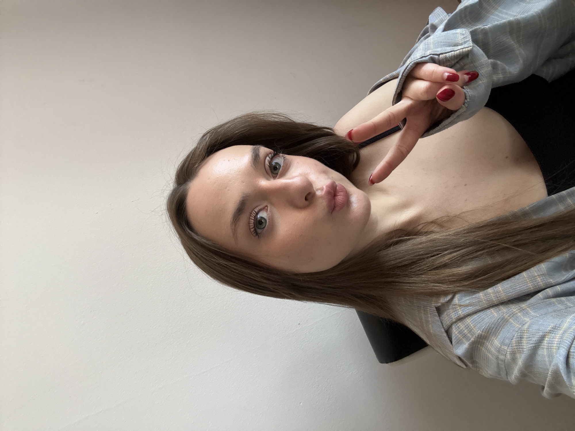 A young woman is depicted in a selfie-style image. Her hair is long, light brown, and styled straight. She has fair skin, green eyes, and is making a kissy face. Her right hand is raised, displaying a peace sign with red nail polish on her fingernails. She is wearing a blue and white plaid shirt. The background is a plain white wall. The location is not discernible from the image.