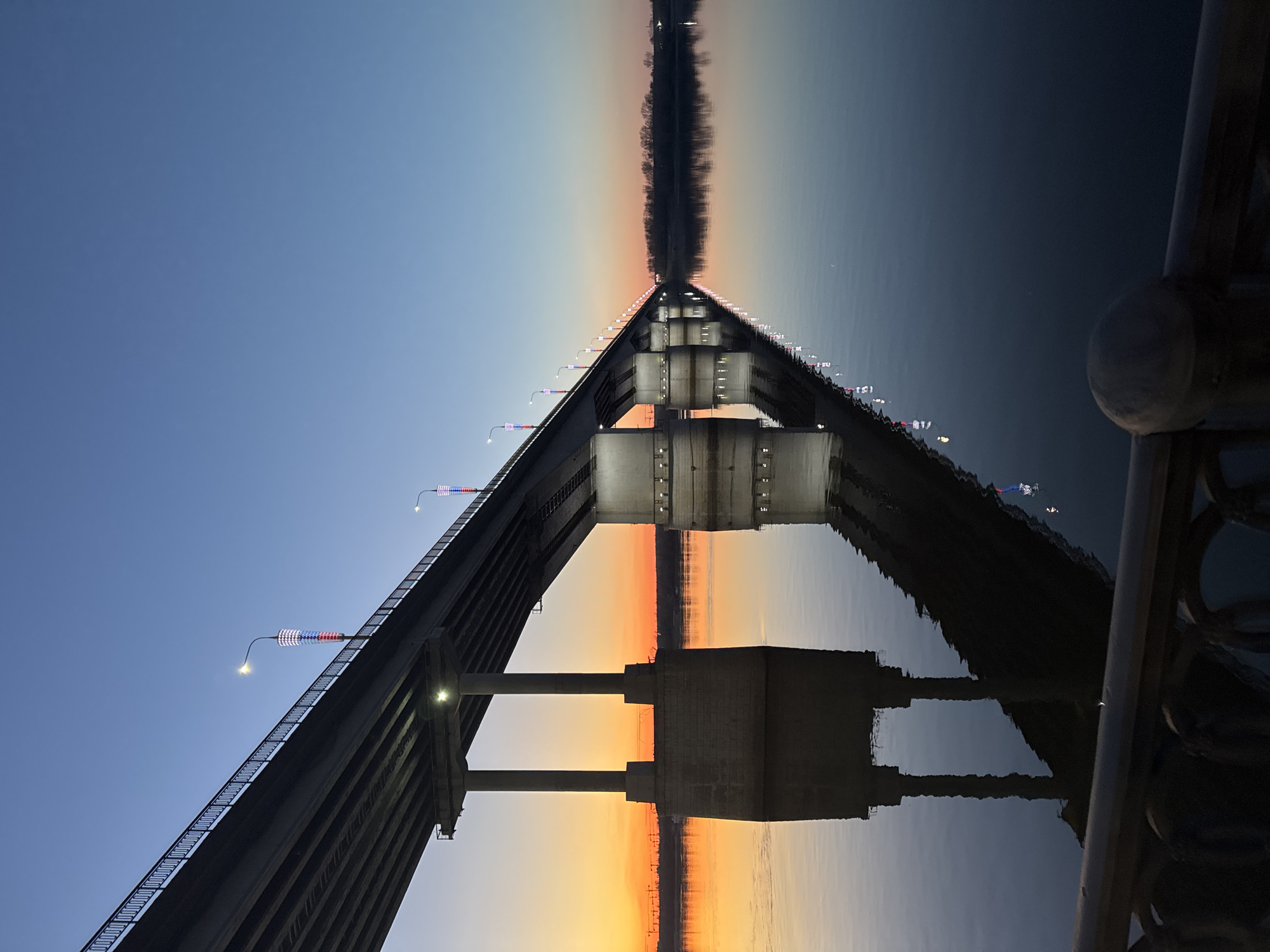 The image depicts a bridge over a body of water at dusk, located in Perm, Russia. The bridge's structure is visible, with multiple supports and illuminated decorative lights along its length. The sky transitions from blue to orange near the horizon, creating a gradient effect. The water's surface reflects the bridge and sky, creating a symmetrical composition. A portion of a railing is visible in the foreground.