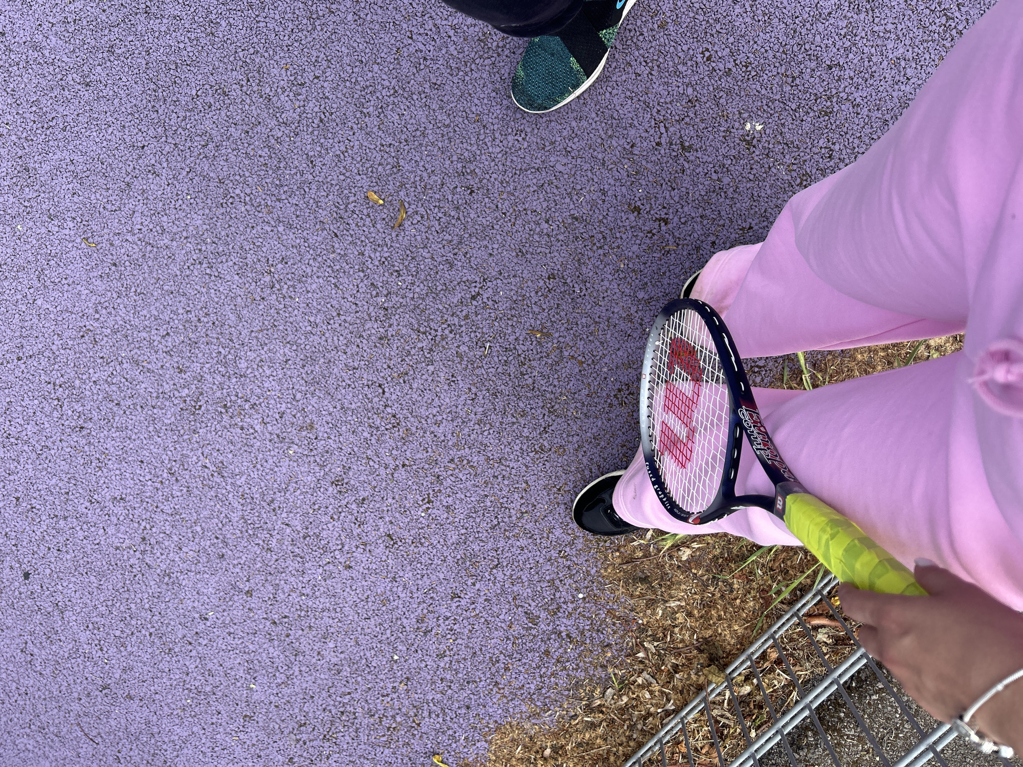 The image shows a person standing on a purple paved surface, possibly a tennis court, holding a tennis racket. The person is wearing light pink sweatpants and black sneakers. Another person's shoe, a green and black sneaker, is visible in the upper part of the image. The lower right corner of the image shows dry leaves, grass, and a metal fence. The location is likely in the city of London, United Kingdom, based on the style of tennis court surfacing used.