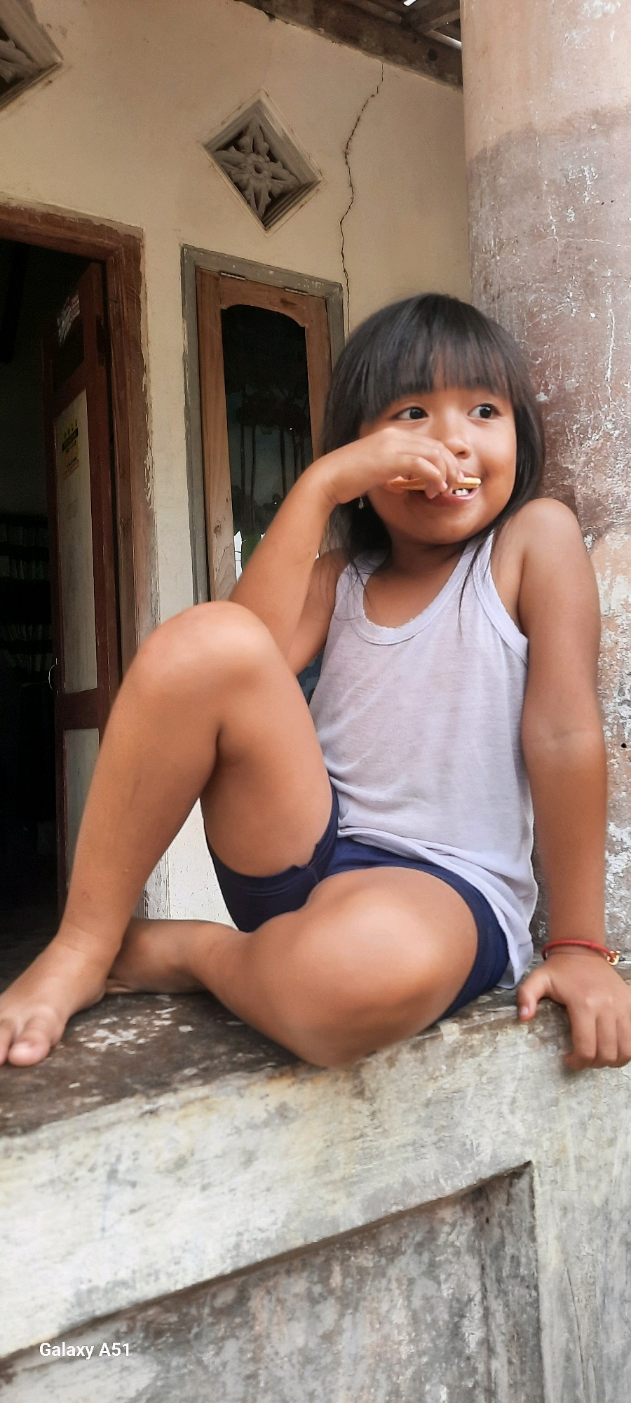 A young girl with dark hair and bangs is sitting on a ledge, eating a snack. She is wearing a white tank top and blue shorts. Her legs are crossed, and her right arm is bent as she brings the snack to her mouth. She is looking to her right with a slight smile. Behind her, a beige building with wooden window frames is visible. A decorative vent is set into the wall above the window. The building appears to be in a rural or semi-rural setting, with a weathered concrete pillar to the right of the girl. A red bracelet is visible on her left wrist. The image has a casual, candid feel, suggesting it was taken during the day. The visible text in the lower left corner reads "Galaxy A51".