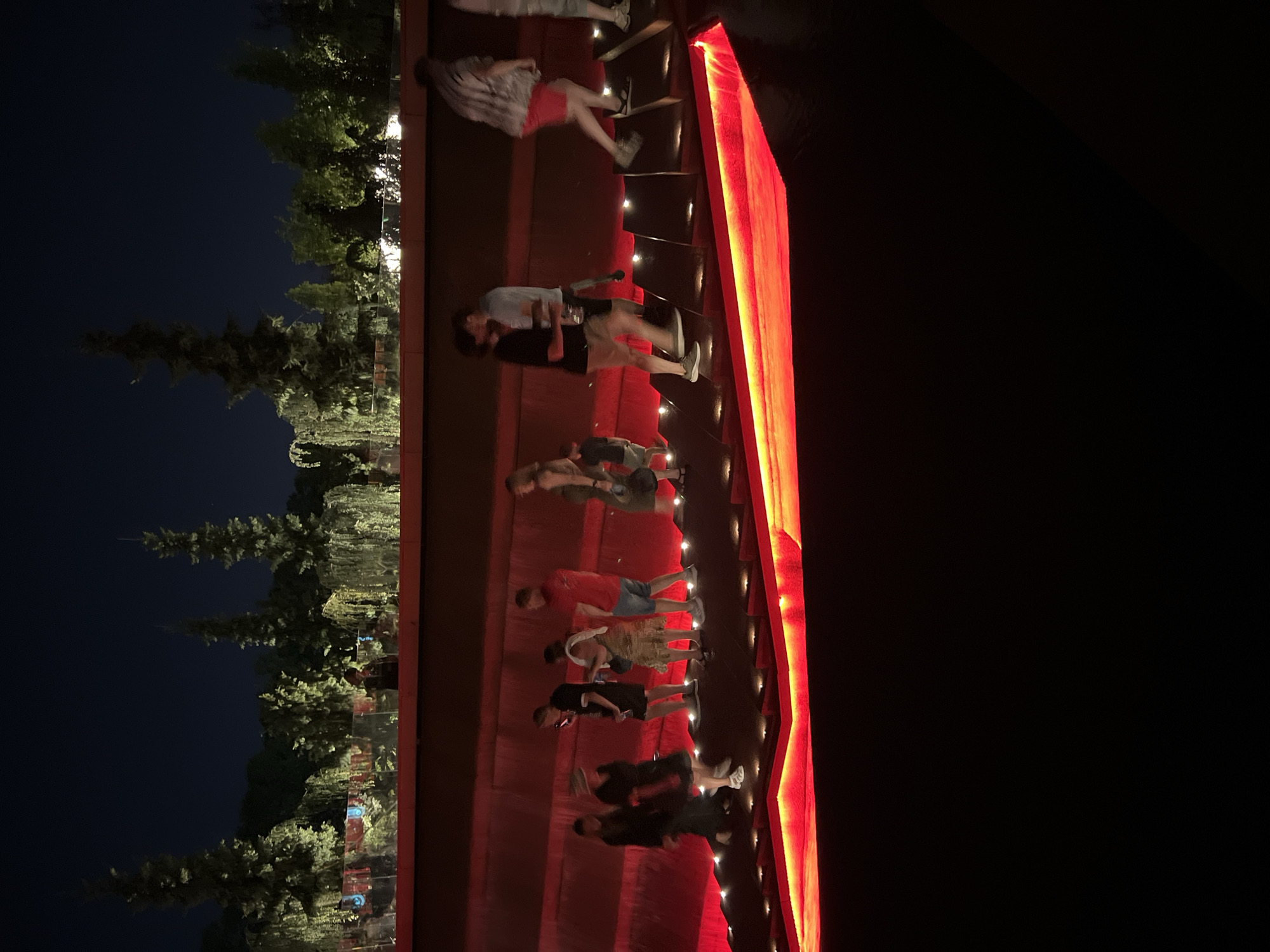 At night, a group of people walk up a series of steps illuminated by red lights, situated within a landscaped area of Krasnodar, Russia. The steps appear to be part of a decorative water feature or a tiered seating area, with the red light creating a vibrant glow. In the background, lush trees and foliage are visible, also lit by artificial lights. The overall scene suggests an outdoor public space designed for evening enjoyment, possibly a park or entertainment venue. The individuals are dressed in casual attire, and their movements indicate they are ascending the illuminated steps, some in pairs and others individually.