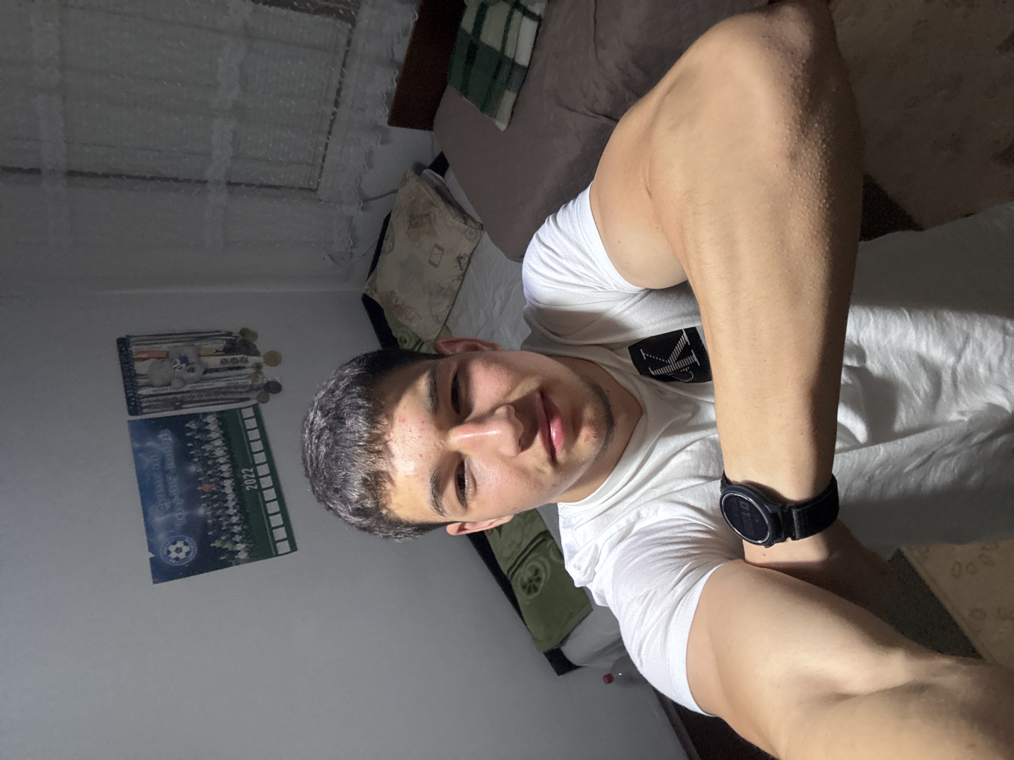 A young man poses for a selfie in what appears to be a bedroom. He's wearing a white t-shirt and a black wristwatch. Behind him is a bed with pillows and a duvet. On the wall, there is a framed picture, possibly a team photo, dated 2022. The location is likely indoors and may be in the city of Zlatograd, Bulgaria, based on external knowledge.