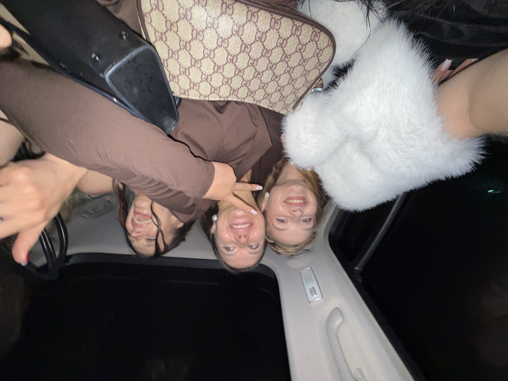 Three females are in the back seat of a car. The woman on the left has dark hair, the middle woman has brown hair, and the right woman has blond hair. All three are smiling. The interior of the car is dark with a white trim. A black purse, a brown Gucci bag, and a white fur-trimmed jacket are visible in the shot. There is no discernible information that would indicate they are located in Belgrade, Serbia.
