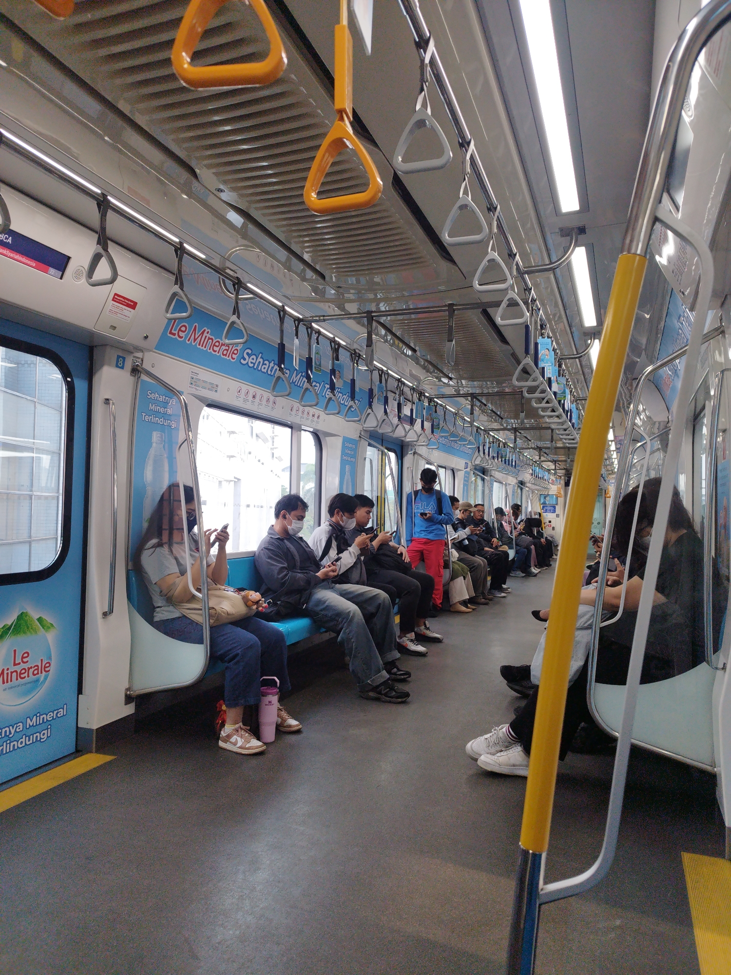 This is a medium-angle, interior shot of a commuter train in South Jakarta, Indonesia. The train car is clean and modern, with blue seats and white walls adorned with advertisements. Several passengers are seated and standing, many wearing face masks. Some are looking at their phones, while others are holding onto the overhead handrails. The lighting is bright, with fluorescent lights running along the ceiling. The overall impression is one of a typical commute in a busy urban environment.