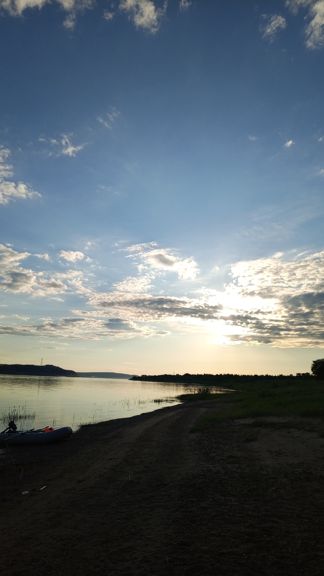 This image captures a wide outdoor landscape scene at either sunrise or sunset over a body of water in Шолья, Russia. The sky occupies the upper two-thirds of the frame, displaying a gradient from deep blue at the zenith to lighter blue and yellow hues near the horizon where the sun is positioned on the right. Numerous white and grey scattered cumulus clouds are visible, particularly concentrated around the sun, which appears as a bright, partially obscured orb casting a radiant glare.
Below the sky, a calm body of water, likely a river or large lake, stretches across the middle ground, reflecting muted tones of the sky. The distant left bank features a dark, undulating landmass with a visible transmission tower or similar utility structure on its summit. The distant right bank is lined with dense, dark green foliage, possibly trees or bushes.
The foreground consists of a sandy or dirt shoreline, appearing dark due to being in shadow relative to the sun's position. A clear, unpaved track or path runs along this sandy bank. On the left side of the foreground, an inflatable boat, grey in color, is beached on the sand. An orange object, possibly a life vest or a piece of equipment, is visible within the boat. Sparse, tall green vegetation grows near the water's edge in the immediate foreground, particularly on the left. No individuals are visible in the scene. Small, dark specks, possibly birds or insects, are discernible in the sky.