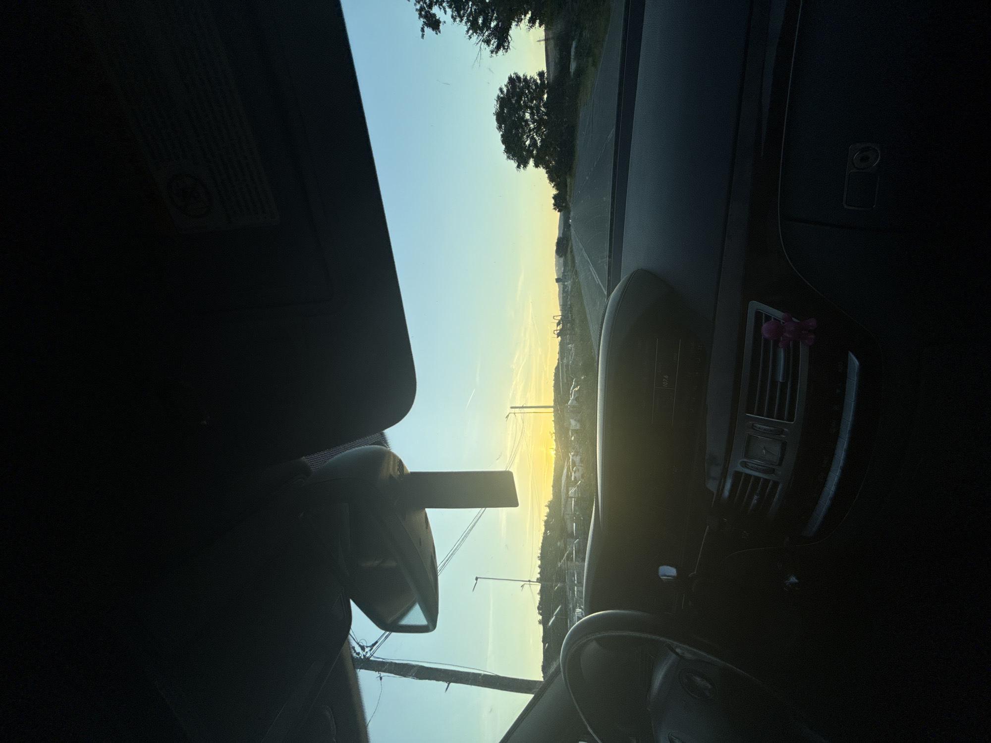 The image depicts a view from inside a car, looking out towards a sunrise. The scene includes elements of the car interior, such as the rearview mirror, sun visor, dashboard, steering wheel, and central console. The exterior view shows a road stretching towards the horizon, with trees and overhead power lines visible. The location appears to be near the city of Piperevo, Bulgaria, judging by the landscape and infrastructure present in the image. No people are visible.
