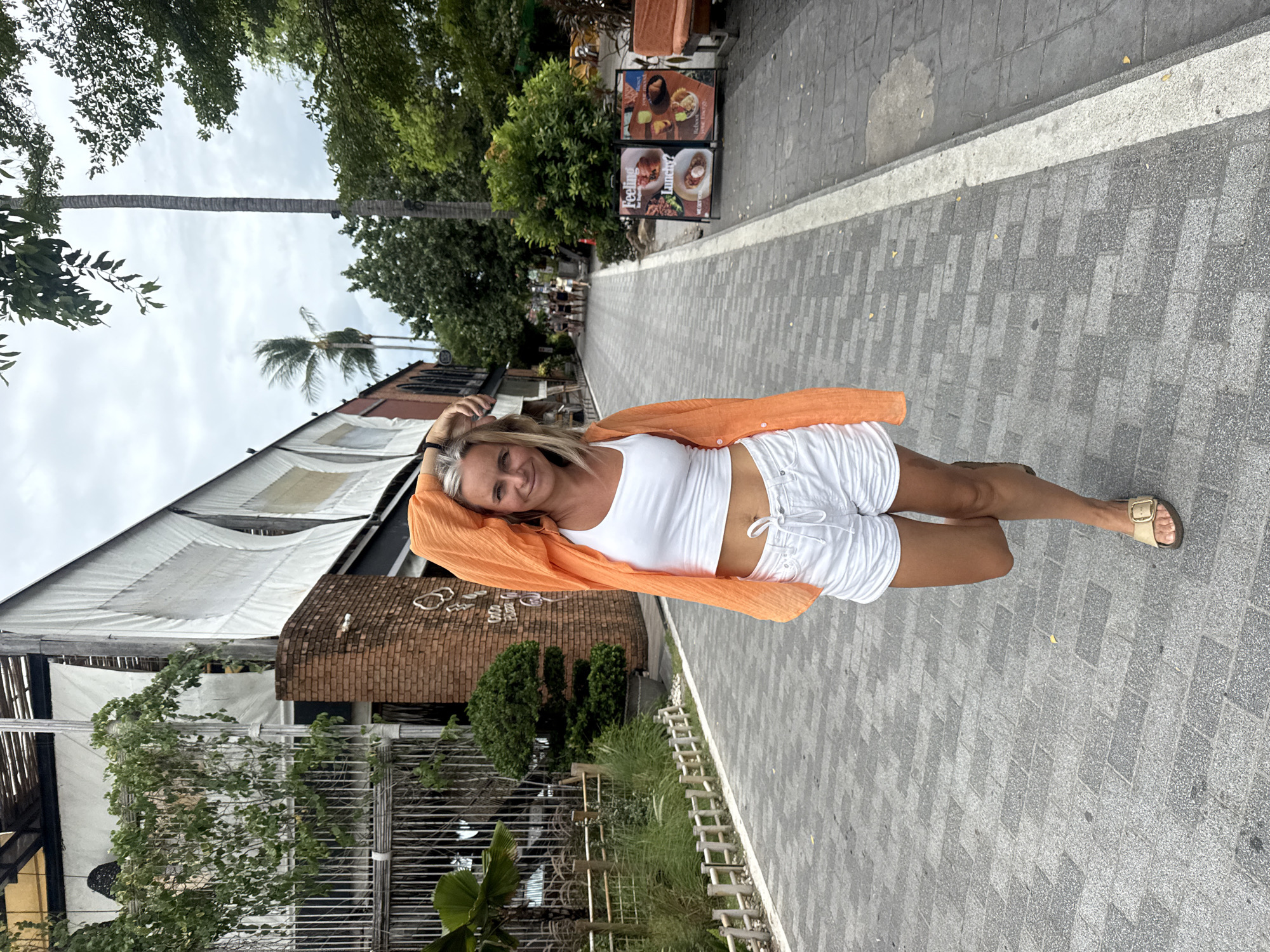 A blonde woman with her arm raised and hand on her head smiles at the camera as she walks down a paved pedestrian walkway in the city of Ko Samui District, Thailand. She is wearing a white crop top, white shorts, and tan sandals. She is also wearing an orange open cardigan over her shoulders. The walkway is lined with trees and buildings. One building on the left has a brick facade and a white awning. On the right, there are signs advertising food and a bench. The sky is cloudy.