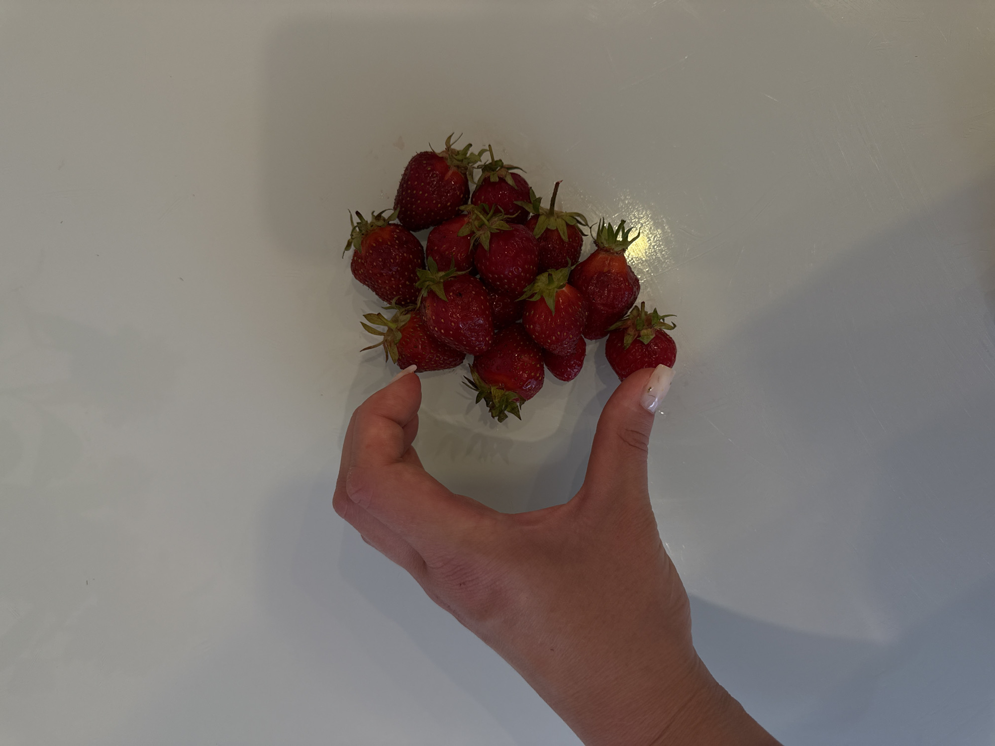 A close-up, top-down shot shows a human hand forming a heart shape, cradling a pile of ripe red strawberries. The hand, with its skin tone and manicured nails visible, is positioned on the left side of the frame, its thumb and index finger creating the curve of the heart. The strawberries, plump and glossy, fill the space within the heart shape and spill slightly out of it. The background is a plain, light-colored surface, likely a table, with subtle shadows suggesting overhead lighting. There are no discernible features that would indicate a specific location within the city of Vladimir, Russia.