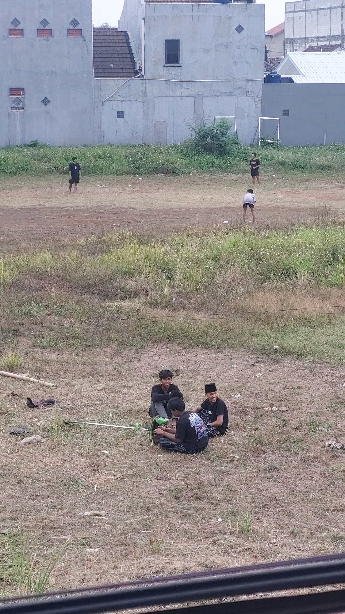 The image captures a group of young males sitting on dry grass in an open field in Bojong Gede, Indonesia. In the foreground, three individuals are seated, with one holding a string trimmer. One of the seated individuals is wearing a black peci, a traditional cap. In the background, two other individuals are standing in the field. Behind the field, a cluster of low-rise buildings with light-colored walls and tile roofs are visible. The overall scene depicts a casual outdoor gathering in a semi-urban environment.