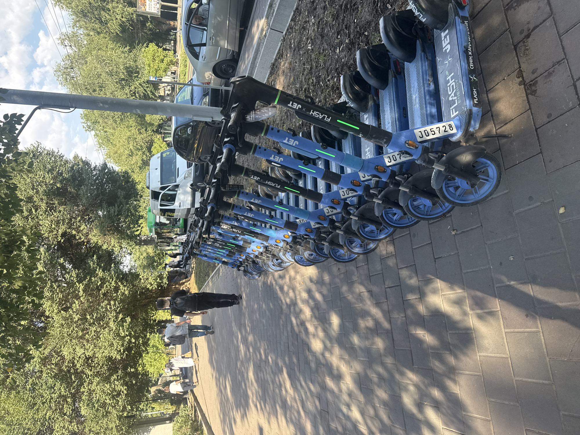 A line of electric scooters, branded "FLASH JET", are parked along a sidewalk in Almaty, Kazakhstan. The scooters are blue and black, with green lights indicating their operational status. Several people are walking on the sidewalk in the background, including two individuals with backpacks. A bus and several cars are visible on the street beyond the scooters. The scene is illuminated by sunlight, casting shadows on the sidewalk. The environment appears to be an urban area with trees lining the street.