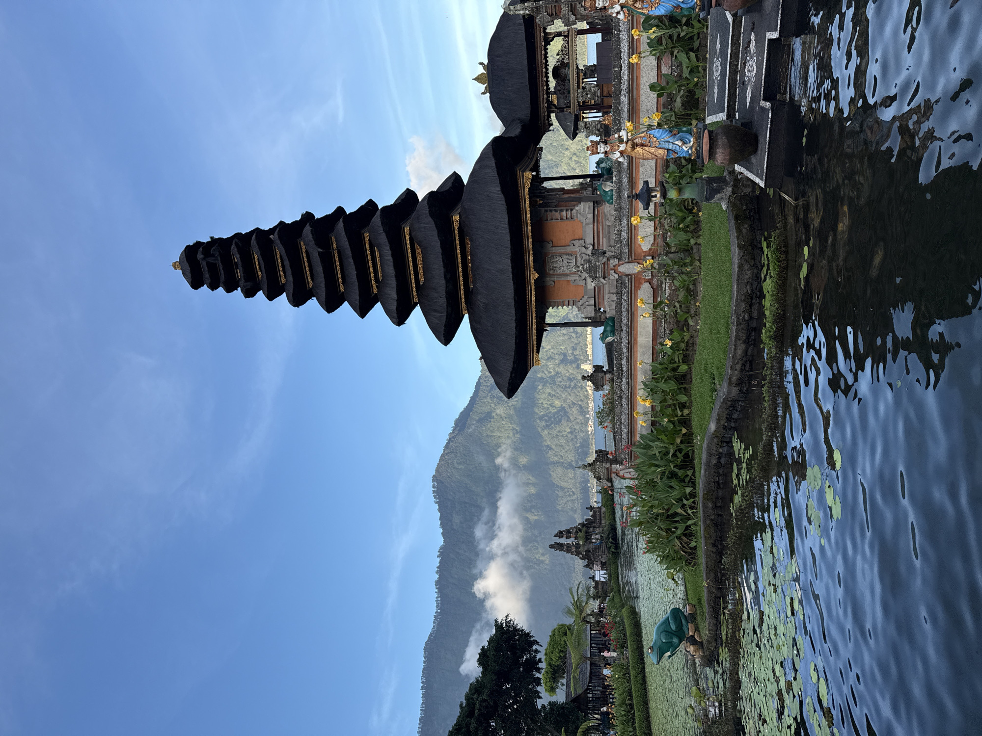 The image depicts Ulun Danu Beratan Temple, a major Shivaite Hindu temple in Bali, Indonesia. It is situated on the edge of Lake Beratan in the mountains near Bedugul, in the city of Baturiti. The temple is a multi-tiered pagoda structure with a distinct tiered roof, constructed from dark materials trimmed with golden ornamentation. Part of the temple's base is submerged in the lake. In the background, a large mountain, partially shrouded in clouds, is visible under a blue sky streaked with light clouds. Lily pads float near the bank of the lake. A green frog statue is seen at the water's edge. Statues and potted plants adorn the landscape near the temple. The orientation of the image is sideways, with the ground and temple on the right side of the frame.