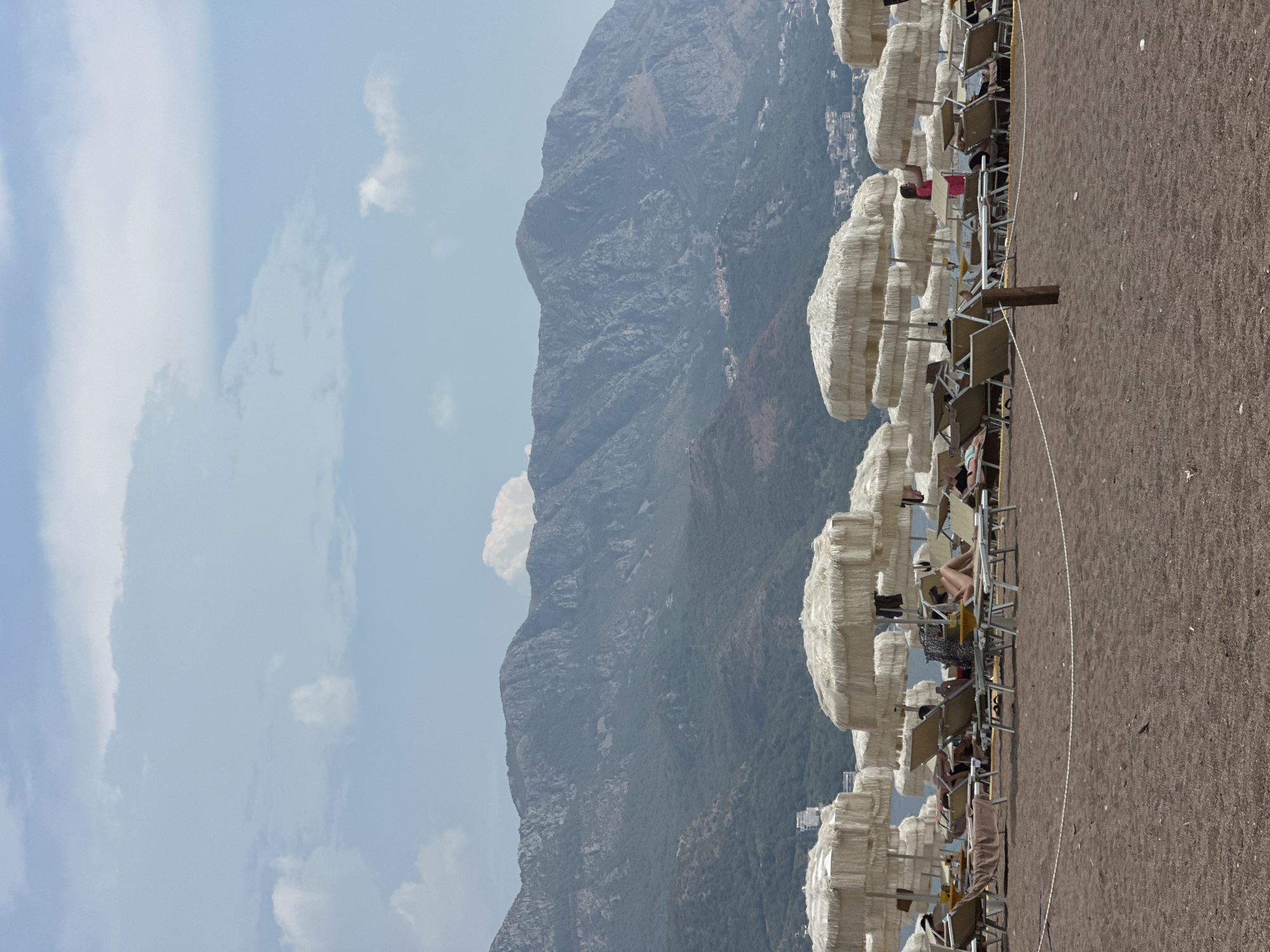 The image depicts a beach scene at Boreti, Montenegro, with numerous beach chairs and umbrellas set up on a dark sand beach. The umbrellas are light-colored and fringed. People are seated on the chairs, some appear to be relaxing or reading. In the background, a range of green, mountainous terrain rises against a partly cloudy blue sky. The sea is visible behind the beach, stretching towards the distant mountains.
