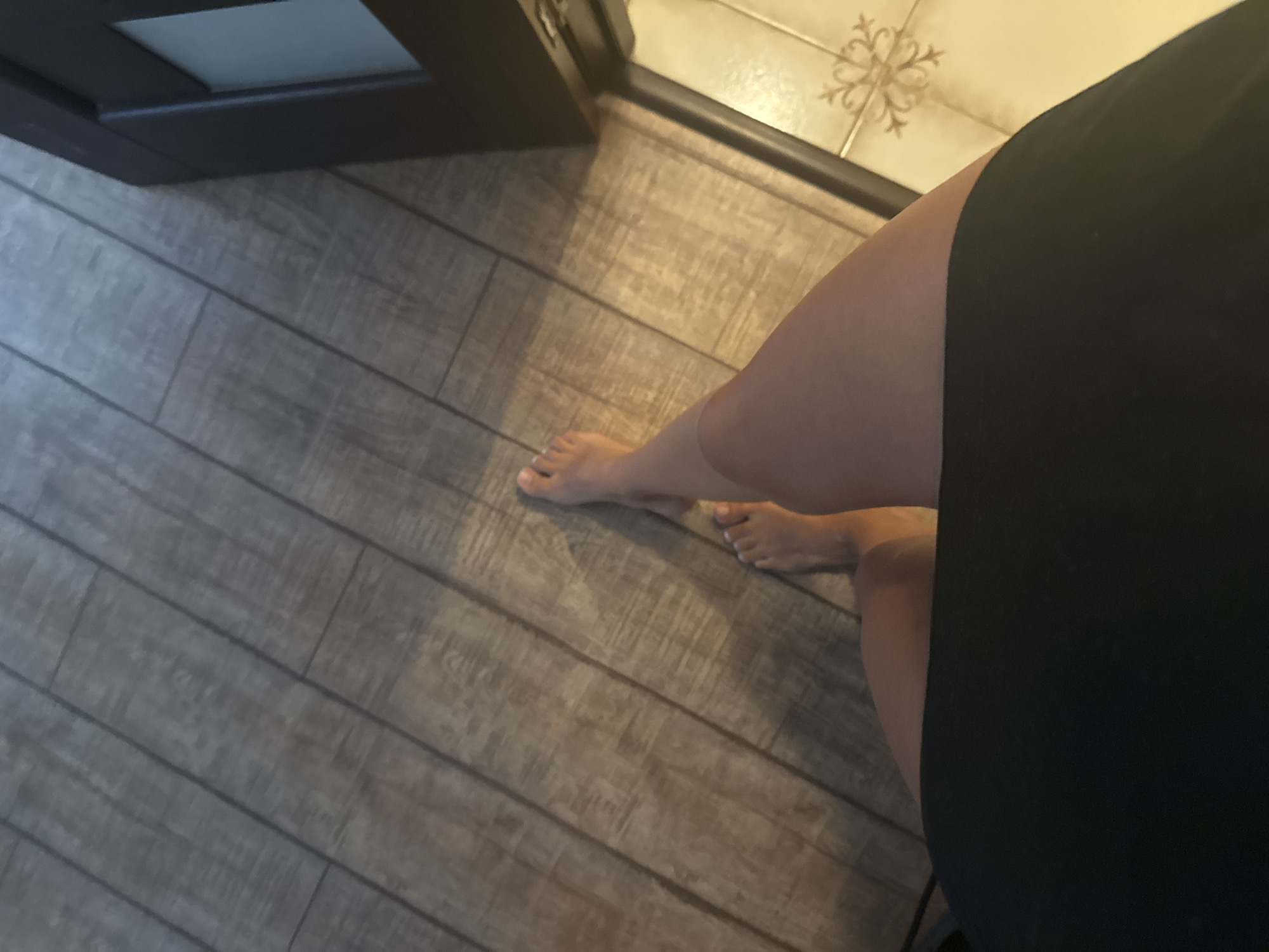 The image shows a person's legs and feet standing on a wood-plank floor. The person is wearing a black skirt or dress. To the right of the frame, a dark brown door frame and a section of a tiled floor are visible. The tiled floor has a decorative pattern in a lighter color. The image is taken from a high angle, looking down at the person's legs. The environment appears to be indoors, possibly a home or a public building. There are no discernible features that would help pinpoint a specific location in Chișinău, Moldova.