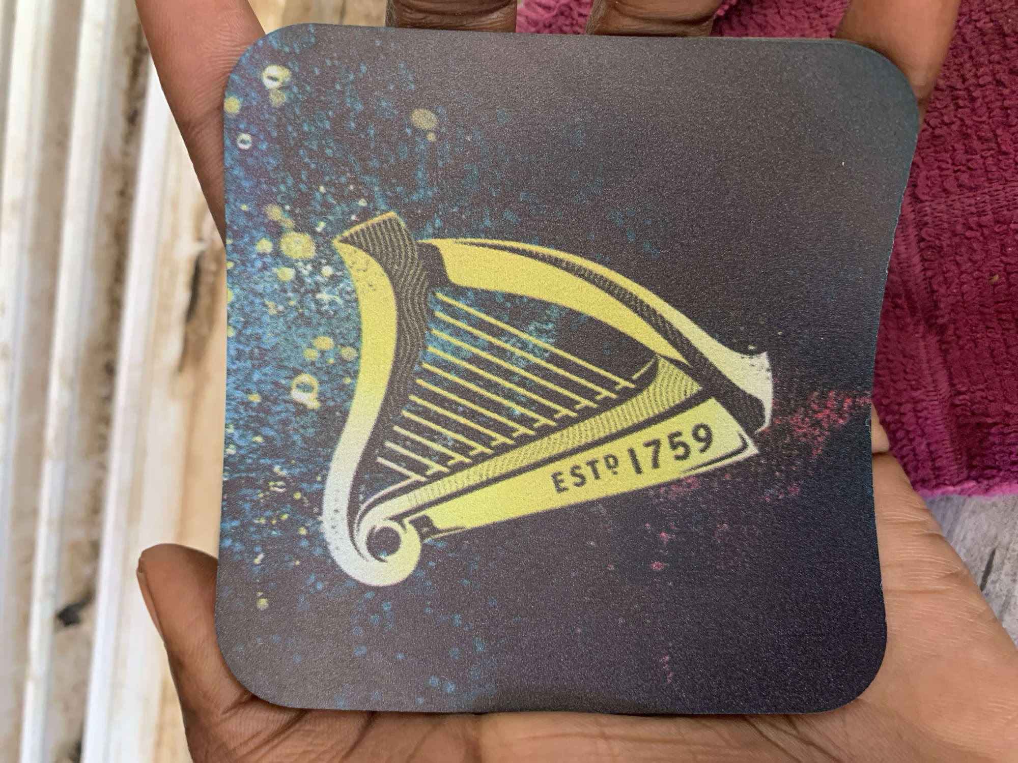 A person is holding a square coaster with rounded corners. The coaster has a dark background with a speckled design in blue, yellow, and white. The logo is a yellow and white harp with "ESTD 1759" printed on a banner-like section of the harp. The person's hands are dark-skinned. Part of a white, ribbed surface is visible on the left side of the image, and a maroon, textured fabric is visible on the right. The location is not discernible from the image. There is no indication of the location being New Stadium, Nigeria.