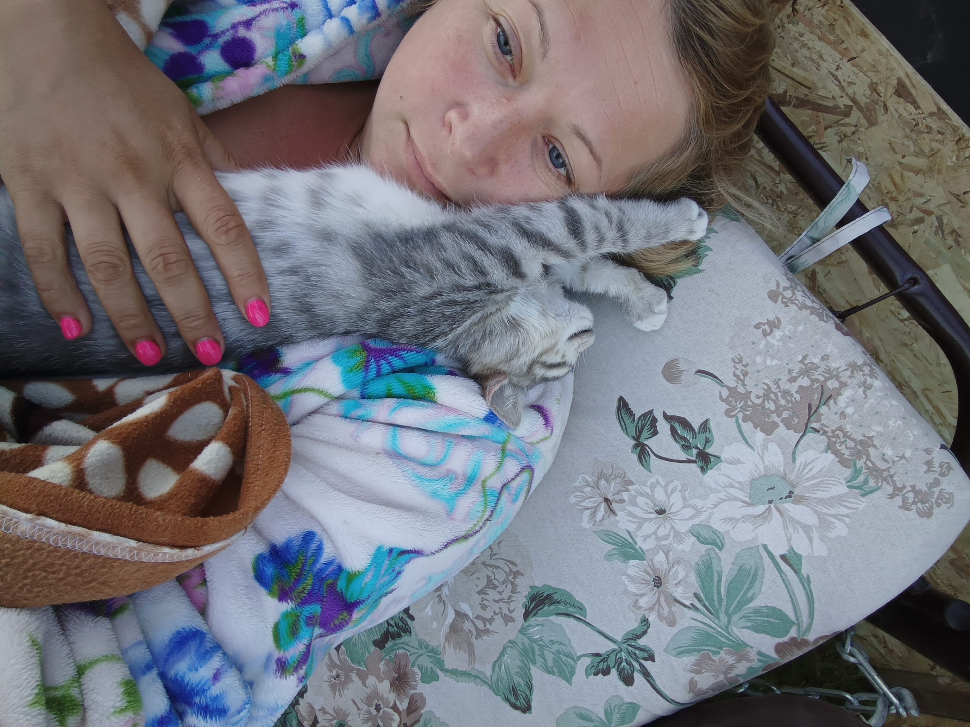 A close-up, high-angle shot captures a person lying down with a cat. The person, a woman with blonde hair and freckles, is positioned on the left side of the frame, her head tilted back and eyes open, looking towards the camera with a relaxed expression. Her hand, with bright pink nail polish, rests on the cat's body. The cat, a grey and white tabby, is curled up comfortably on the woman's chest, its head resting on a floral-patterned pillow. The woman is wrapped in a brightly colored, patterned blanket, which is partially visible. The background consists of a wooden surface and a dark metal structure, suggesting an outdoor setting, possibly in a park or a garden in Ivanovskoe, Russia, given the context of previous images. The overall impression is one of peaceful intimacy and comfort between the woman and her pet.
