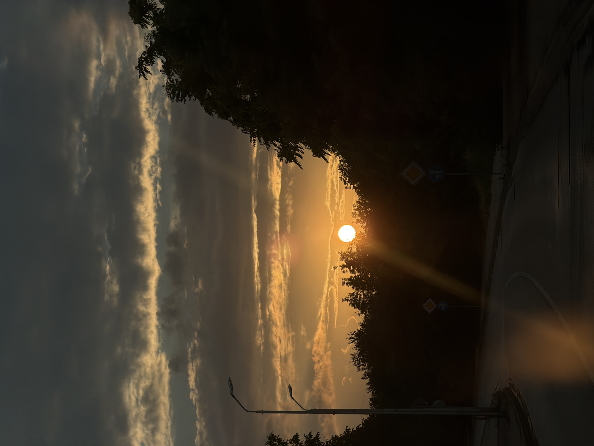 The image captures a road in Lovech, Bulgaria, during a sunset. The sun is partially obscured by dark clouds, casting an orange glow through them and creating visible sunbeams. The foreground shows a paved road with a white line, reflecting the sunset's light. On the left side of the road, there are two streetlights and a sidewalk. On the right side, a dense line of trees is silhouetted against the bright sky. Two road signs are visible on posts along the roadside: one diamond-shaped sign and a circular blue sign below it. The overall atmosphere is serene and twilight-like.