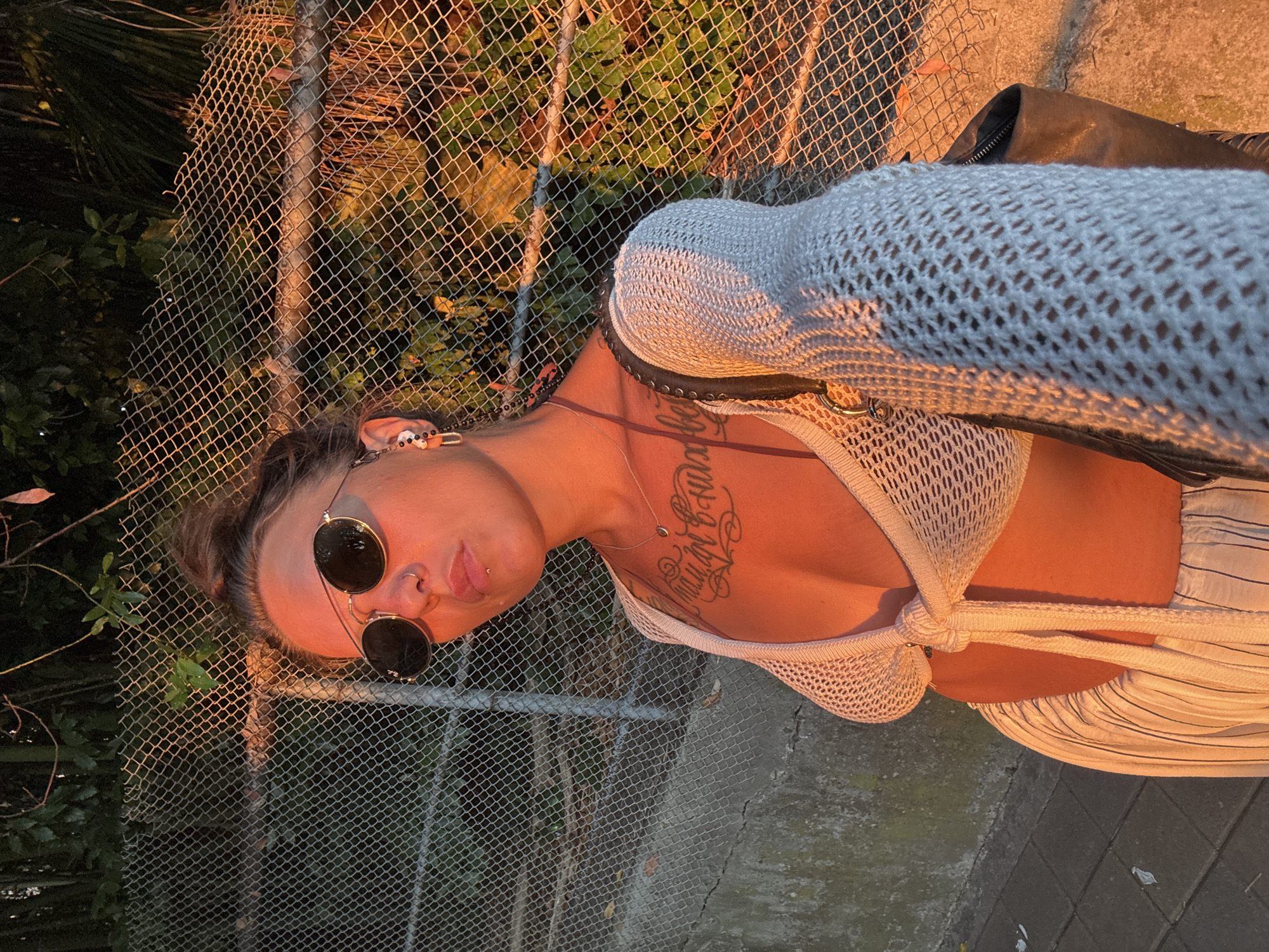A young woman stands in front of a chain-link fence, bathed in the warm glow of sunset. She is wearing a white, crocheted halter top that ties at the front, exposing her chest adorned with a tattoo. Her hair is pulled up in a messy bun, and she wears aviator sunglasses and hoop earrings. A silver necklace with a small pendant rests on her collarbone. She is also wearing striped pants. The background consists of the chain-link fence, some greenery, and a concrete wall. The lighting suggests it is late afternoon or early evening. There are no specific indicators in the image that place the location in Sochi, Russia.