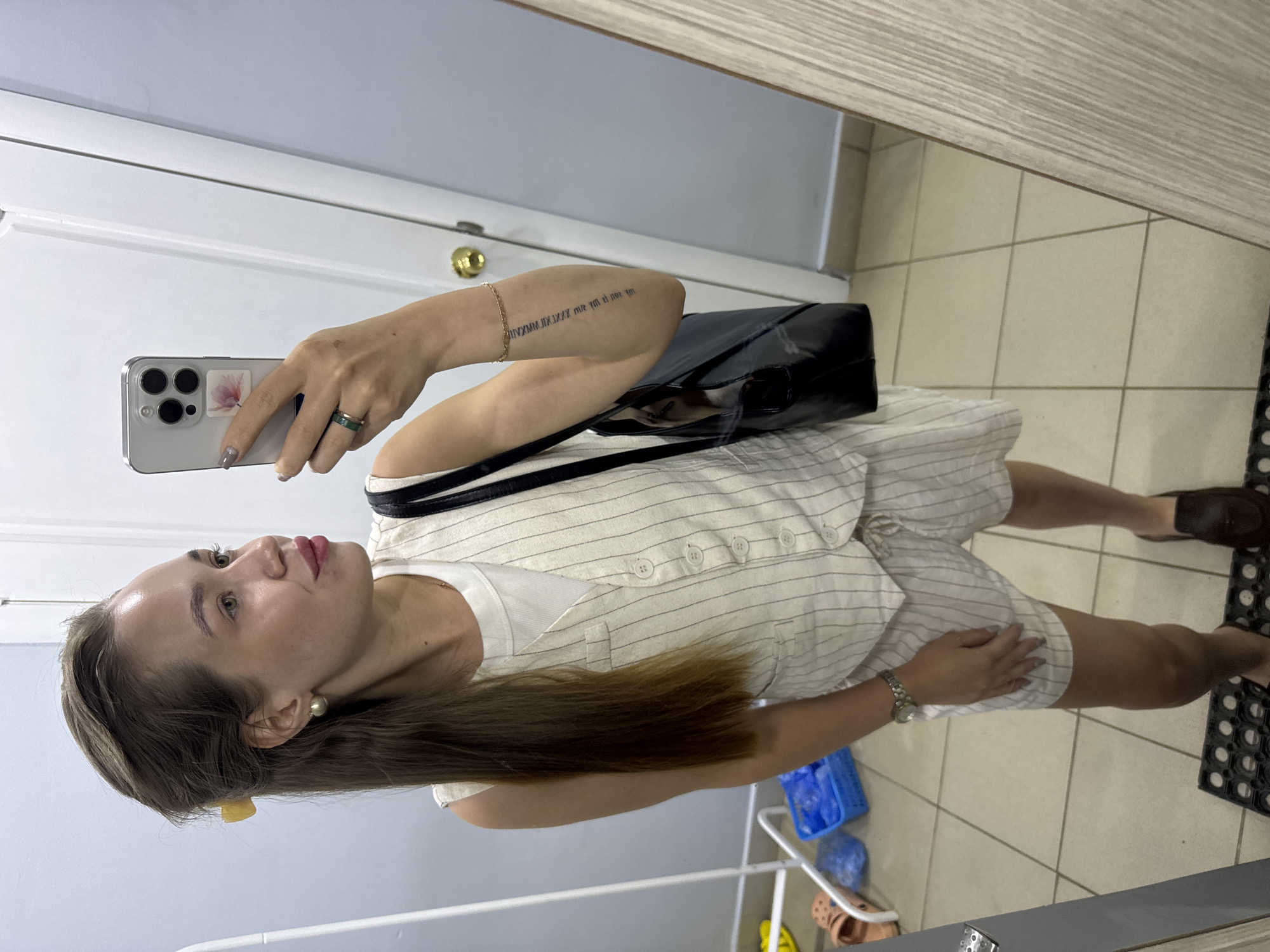 A woman is taking a selfie in a bathroom with tiled floors and white walls. She is wearing a light-colored striped romper with a white top underneath, a ring on her finger, and a watch on her wrist. She is carrying a black purse over her shoulder. Her hair is brown and pulled back with a yellow clip. The bathroom contains a metal towel rack and various cleaning supplies. The image does not confirm that this is the city of Chaikovsky, Russia.