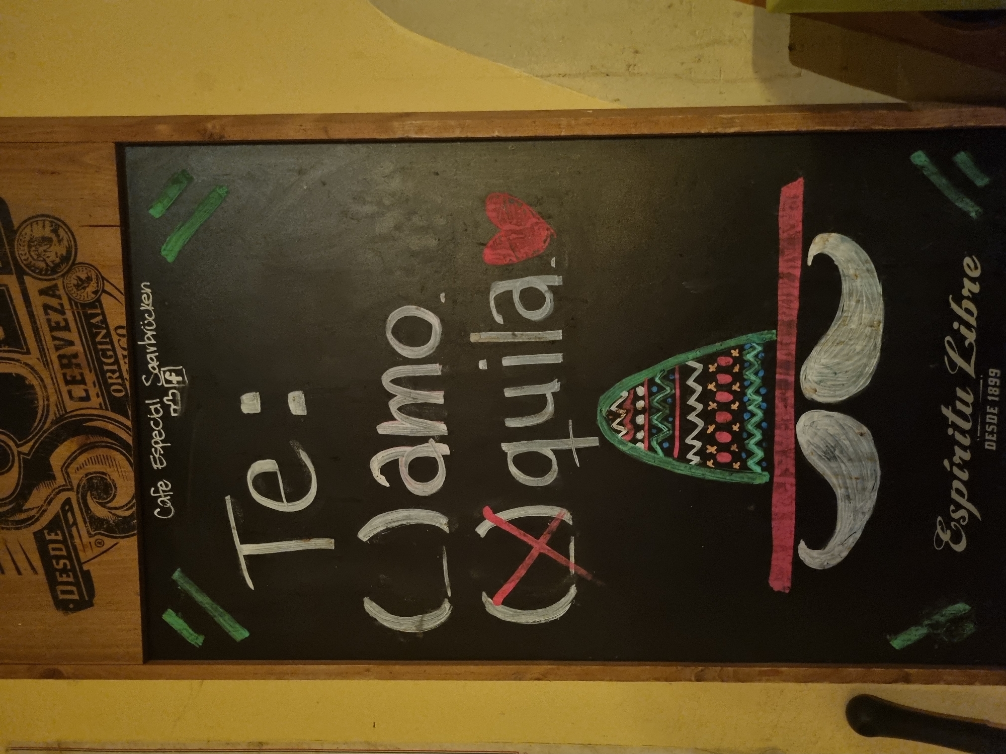 A chalkboard menu, bordered by wood, is hanging on a wall. The menu has writing in chalk, including the words "Te: amo quila," written vertically. A heart shape is above the word "quila," and a large X crosses the word. There is a drawing of a cocktail with decorative designs, a pink line representing the glass stem, and two white curved lines for the moustache of the man drinking. On the left side of the chalkboard is the word "DESDE CERVEZA ORIGINAL," and "Cafe Especial Saarbrücken" is written above the first part of the Spanish words. On the right side, the words "Espiritu Libre DESDE 1899" are written vertically. The context of the menu appears to be a restaurant in Saarbrücken, Germany.
