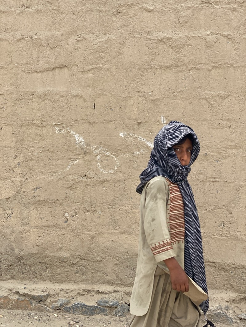 A young person, likely a child or adolescent, stands outdoors against a textured, light brown mud or stucco wall in Kandahar, Afghanistan. The individual wears a light-colored tunic (possibly part of a shalwar kameez) with subtle embroidered patterns in brown and white on the sleeves and side, paired with matching loose-fitting pants. A dark blue or black and white patterned headscarf, possibly a keffiyeh, is draped over their head and shoulders, partially obscuring the lower half of their face but leaving one eye visible. The person holds a light-colored, folded object, possibly paper or fabric, in their right hand.
The gaze is directed towards the viewer, conveying a serious or contemplative expression. The wall behind them shows signs of age and wear, with visible white markings or faded graffiti that appear to be in Arabic or Persian script. The ground in the foreground is unpaved, consisting of dirt and small stones. The lighting suggests daylight, with soft shadows, indicating it could be an overcast day or a specific time of day with indirect sunlight. The overall scene depicts a moment of daily life in an outdoor setting, characterized by traditional attire and architectural elements common to the region.