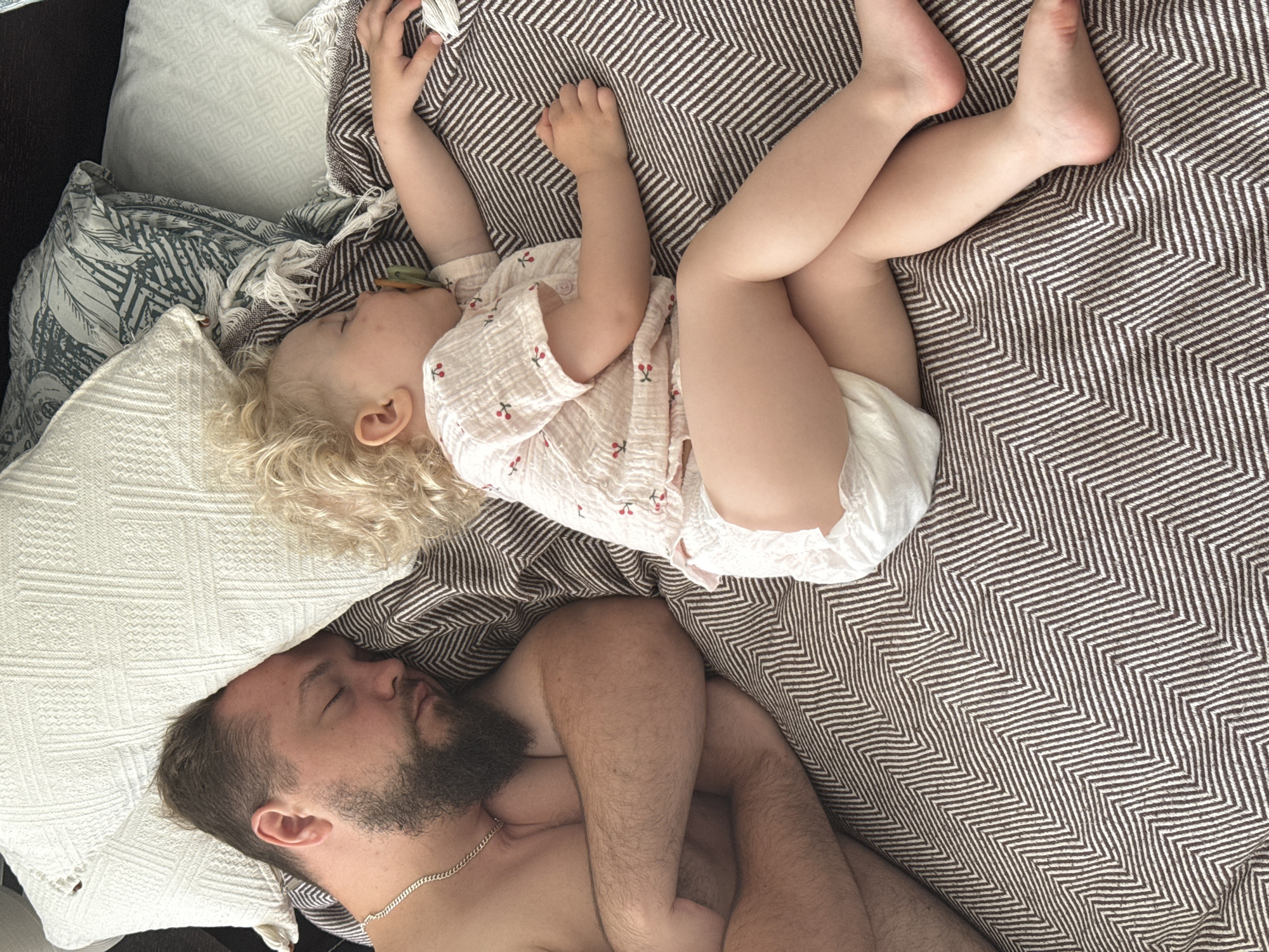 The image shows a man and a young child sleeping on a bed. The man is on the left side of the frame, lying on his back with his arms crossed. He has a beard and is wearing a silver chain. The child is on the right side, lying on their side with their legs crossed. The child has blonde curly hair and is wearing a light-colored shirt with a cherry pattern and a diaper. A pacifier is in the child's mouth. The bed is covered with a brown and white chevron patterned blanket, and there are pillows in the background. The scene appears to be in a domestic setting. The location in the city of Vladimir, Russia, is not ascertainable from the image.