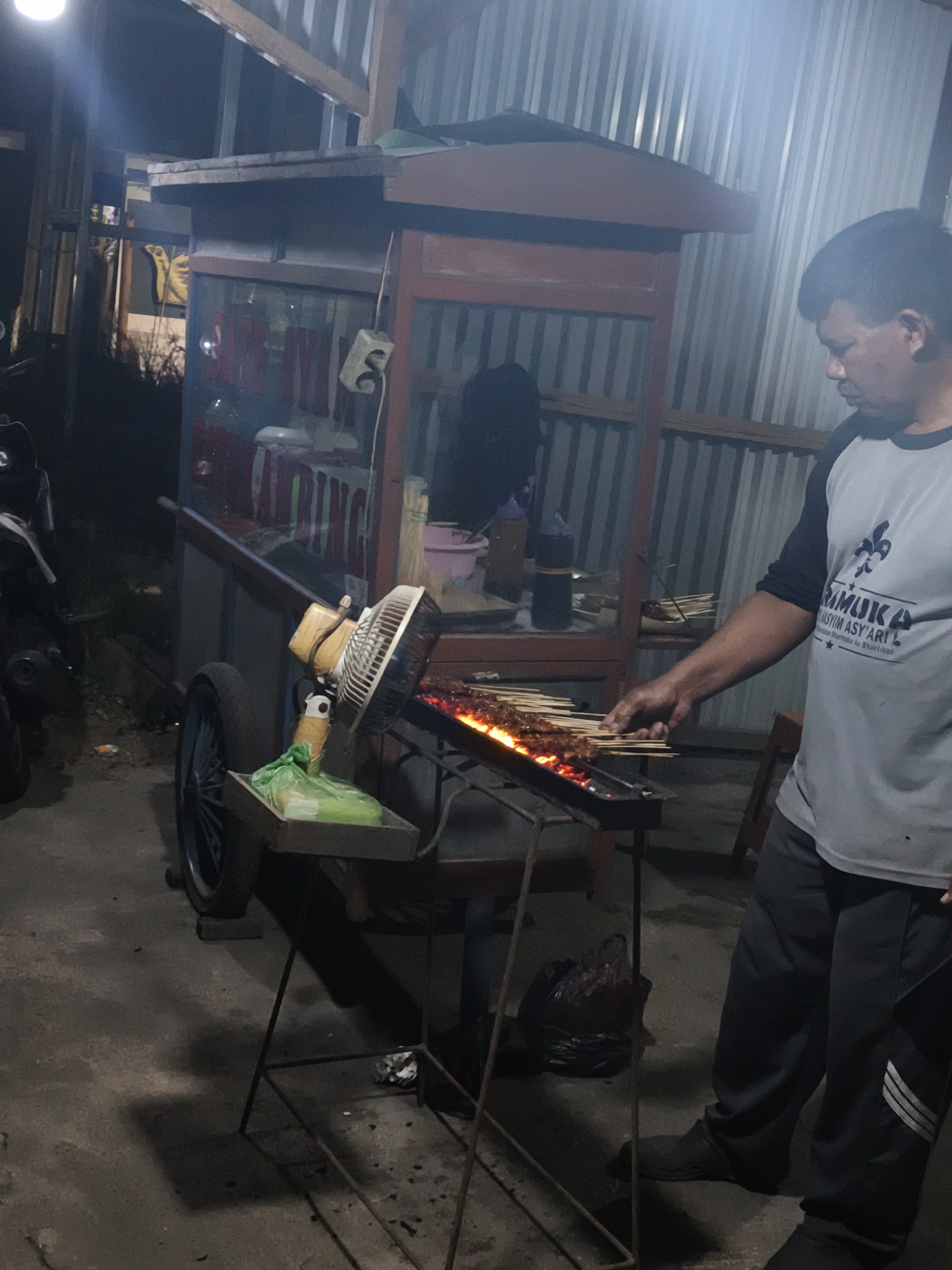 A man is standing next to a food cart, grilling meat skewers over hot coals. The cart is located outdoors, likely on a street in Sidorejo, Indonesia. The man is wearing a grey t-shirt and dark pants. He is holding a metal skewer of meat in his right hand and appears to be turning it over the grill. The food cart is made of metal and wood, with a glass display case and a fan mounted on the side. Several skewers of meat are already on the grill, cooking. The background shows a corrugated metal wall and some other structures, suggesting a street-side vendor setup. The lighting is dim, indicating it is likely nighttime.