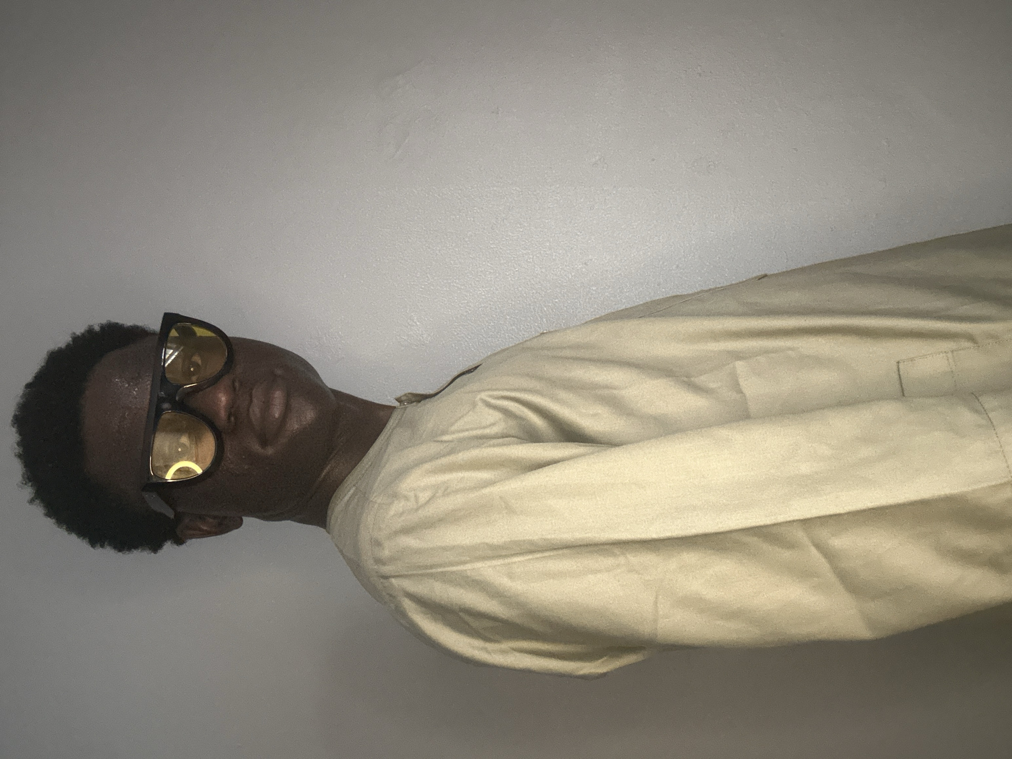 A medium shot shows a young Black man from the chest up, facing right, with his head turned to look at the camera. He has dark, curly hair and is wearing thick-rimmed, square sunglasses with yellow lenses. His expression is neutral, with his mouth slightly open. He is wearing a light tan, long-sleeved garment that appears to be a traditional African robe, possibly an Agbada or Buba, with a subtle texture and what looks like a pocket on the right side of his chest. The garment is unbuttoned at the neck, revealing a hint of his collarbone. The background is a plain, light gray wall with subtle textural variations and faint shadows. The lighting is dim, with a light source casting shadows from his face and casting a yellow reflection on his sunglasses. The scene is likely indoors, possibly a home or studio, and while the individual's attire suggests a Nigerian context, the image itself provides no specific visual cues to pinpoint its location within Badagry, Nigeria.