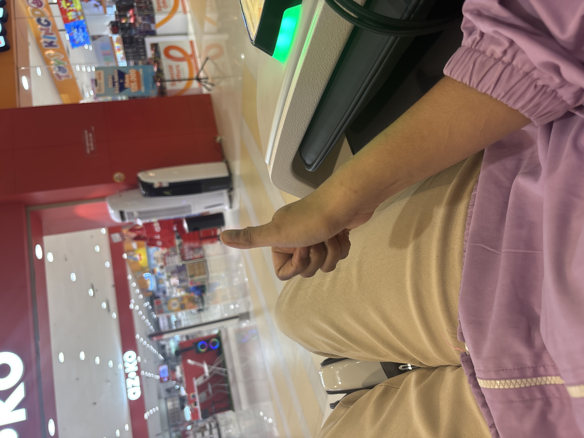 The image shows a first-person perspective of a person in a mall, likely a department store. The person is sitting and giving a thumbs-up gesture. They are wearing tan pants and a purple jacket with a zipper. In the background, the interior of a mall is visible, with store fronts including "AZ.KO" and "TOYS KINGDOM" signs. The floor is shiny and reflective. There are display shelves and what appears to be a massage chair or lounge seating in the foreground. The environment suggests an indoor shopping mall setting in North Jakarta, Indonesia.