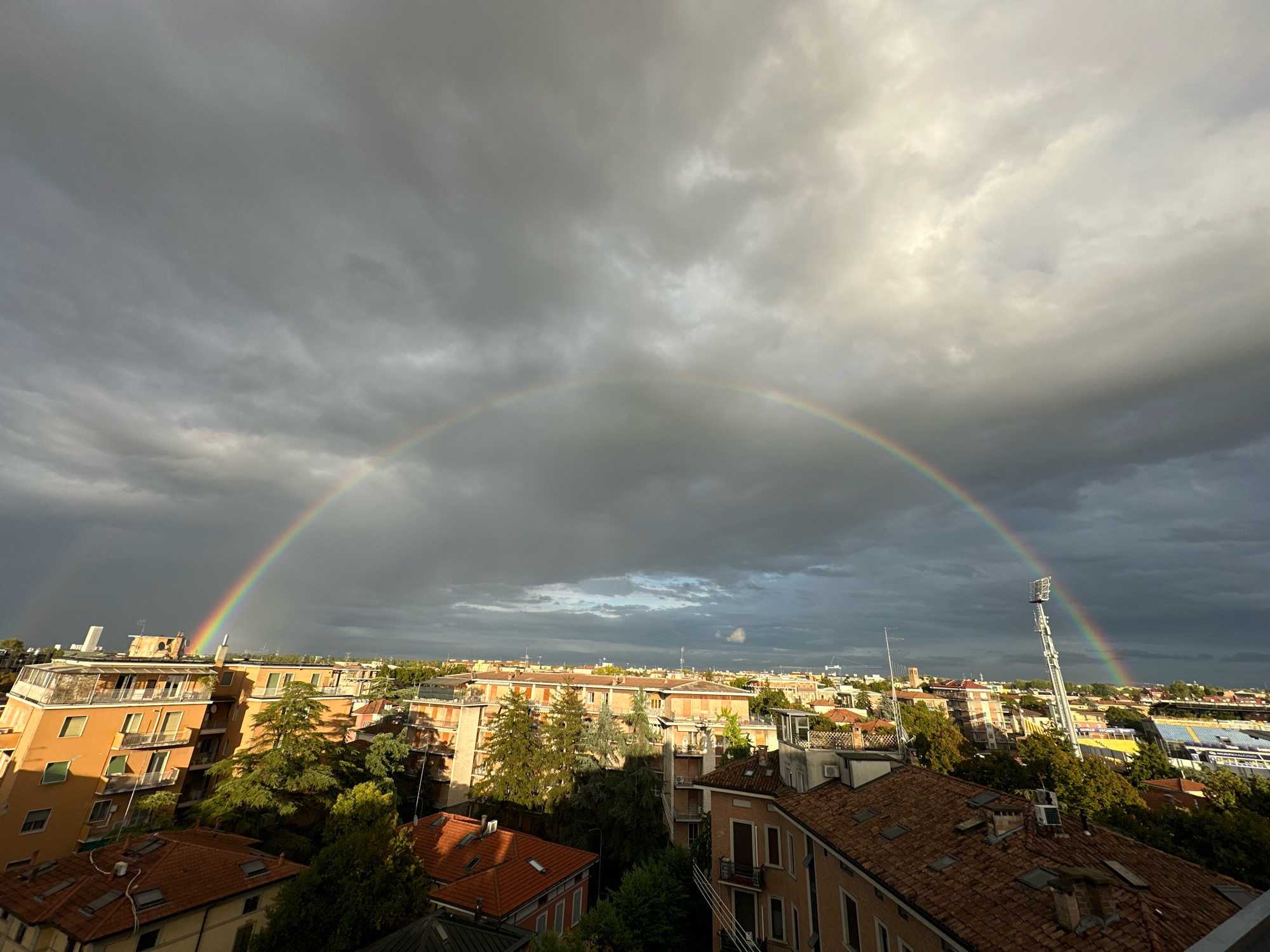 This is an eye-level, wide-angle photograph taken from a high vantage point overlooking a city. The sky is dominated by heavy, dark gray clouds, with patches of brighter, lighter gray and white clouds visible, particularly towards the upper right of the frame where sunlight is breaking through. A prominent, vibrant rainbow arches across the sky, extending from the left side of the image towards the right.
Below the sky is a dense urban landscape. In the foreground and mid-ground, there are numerous buildings with terracotta tiled roofs and orange, beige, and white facades. Many buildings feature balconies with railings, and some have visible windows and air conditioning units. Lush green trees are interspersed throughout the cityscape, some reaching up to the level of the buildings.
On the right side of the image, a tall, silver stadium light tower stands out against the sky. Further in the distance, a blue and yellow striped stadium is visible. The overall impression is of a city skyline under dramatic weather conditions, with the rainbow offering a striking contrast to the brooding clouds. Based on the architecture, particularly the stadium and the style of buildings, this scene is likely in Parma, Italy. There are no people visible in the photograph.