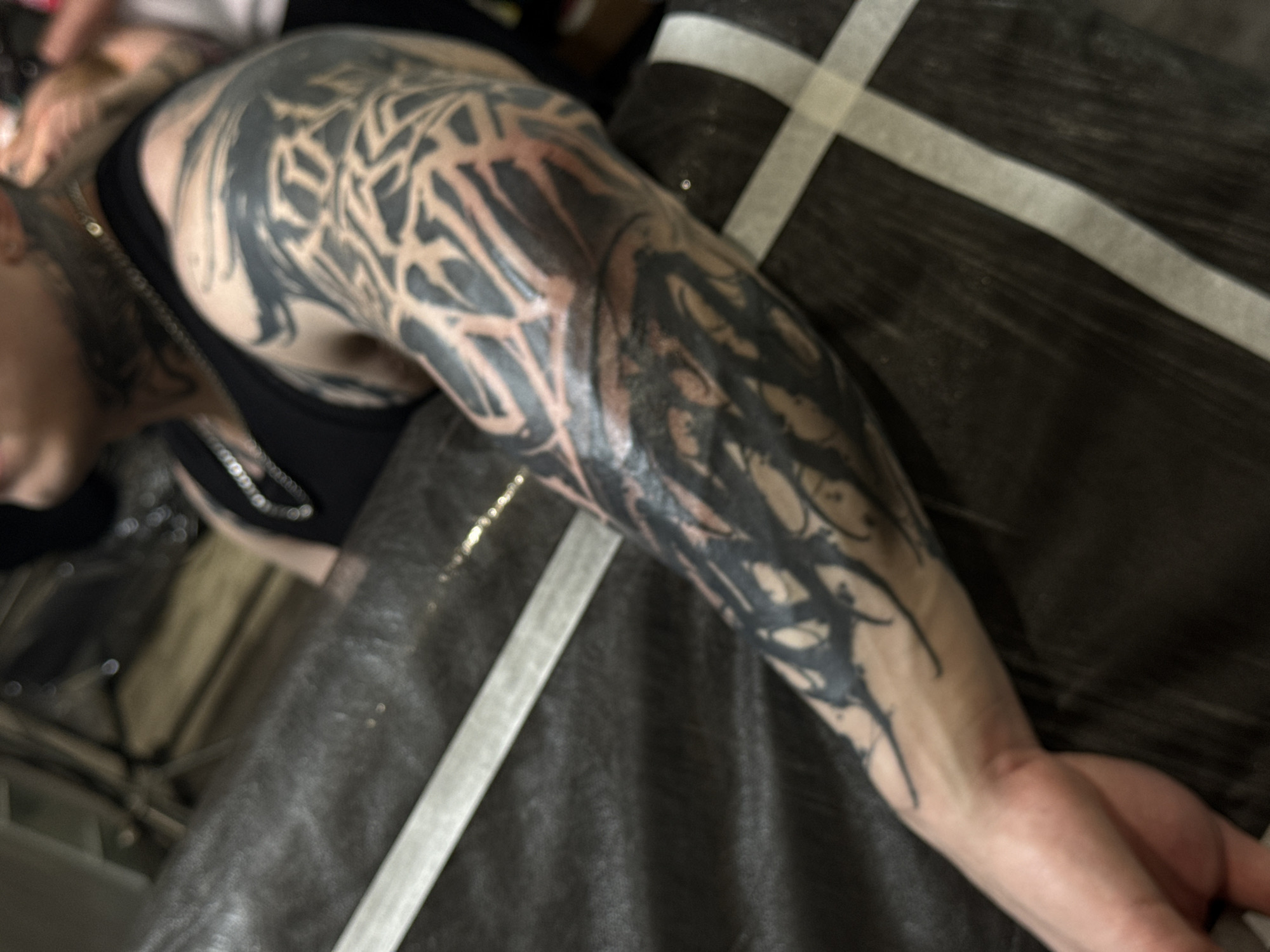 The image shows a person lying down, with their left arm extended and heavily tattooed. The tattoos are primarily black ink and appear to be abstract and tribal in design, covering the shoulder, upper arm, and forearm. The person is wearing a black tank top and a silver chain. The arm is resting on a dark surface with light-colored tape forming a cross pattern. The location is not possible to determine from the image alone.