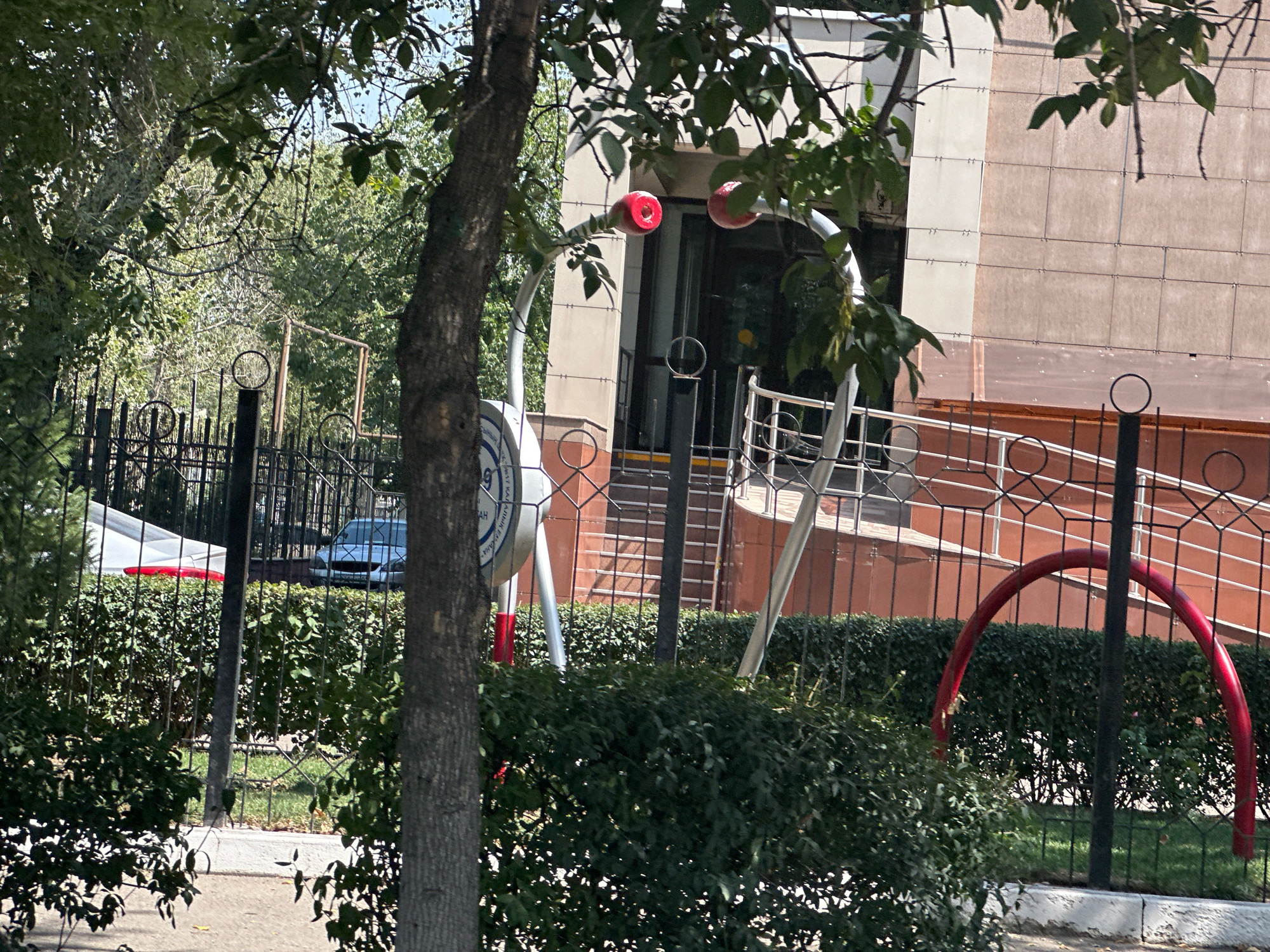 The image is an outdoor, eye-level shot captured on a sunny day in Almaty, Kazakhstan. The scene depicts a building with a textured, beige facade, featuring a darker trim line approximately one-third of the way up from the bottom. To the left of the building, a tree with textured bark and green foliage partially obscures the view. In the foreground, a black metal fence encloses a manicured hedge. Beyond the fence, a light-colored car is visible parked behind the hedge, and further back, more trees and foliage are present. A red arch structure is visible in the middle ground, supported by a black pole. Several circular decorative elements are integrated into the fence posts. The overall setting suggests an urban environment with green spaces.
