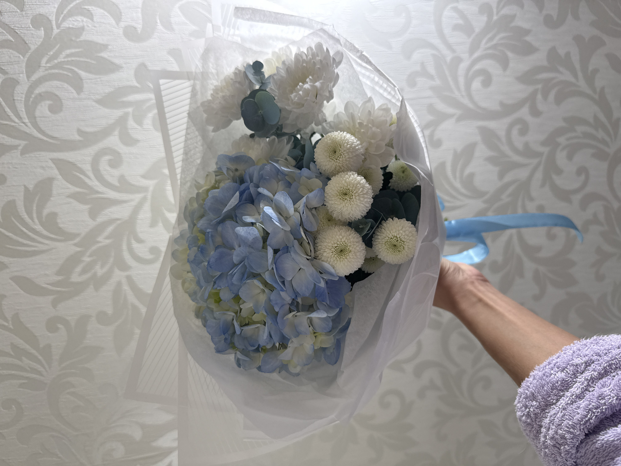 A person in a purple, textured robe holds a bouquet of flowers against a light grey wallpaper with a swirling leaf pattern. The bouquet is wrapped in white paper and tied with a light blue ribbon. It contains blue hydrangeas, white chrysanthemums, and greenery. The location is Unknown, Russia.