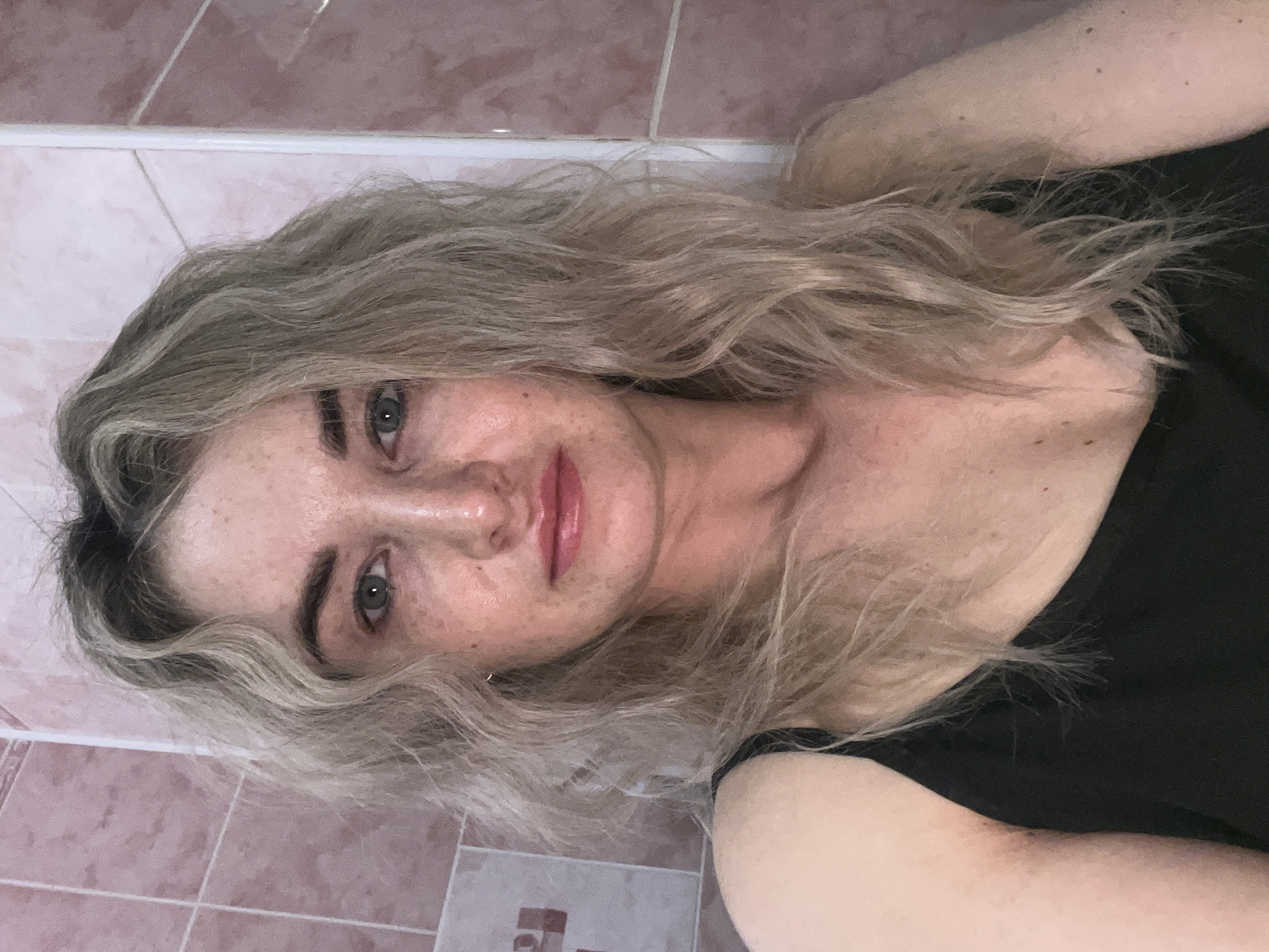 A woman is lying down, her head resting against a tiled wall. She has fair skin with freckles, light eyes, and wavy blonde hair. She is wearing a black top. The background consists of pink and white tiles. The image does not provide sufficient information to verify the location as Chaikovsky, Russia.