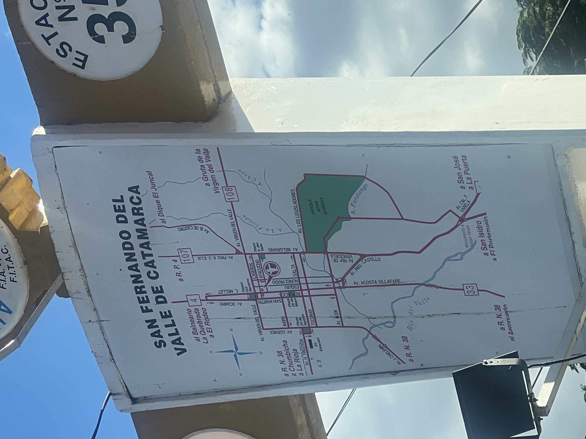 The image shows a sign with a map of San Fernando del Valle de Catamarca, Argentina. The sign is located at an intersection, with roads branching off in multiple directions. The map highlights routes to various locations within the city and surrounding areas, including La Quebrada, El Juncal, Chumbicha, La Rioja, San José, and San Isidro. Key landmarks, such as Parque Adan Quiroga and Plaza 25 de Mayo, are marked on the map. The sign also displays road numbers, such as R.P. 4, R.P. 107, R.P. 108, R.N. 38, and the number 33, indicating highway routes. The overall environment suggests an outdoor setting within the city.