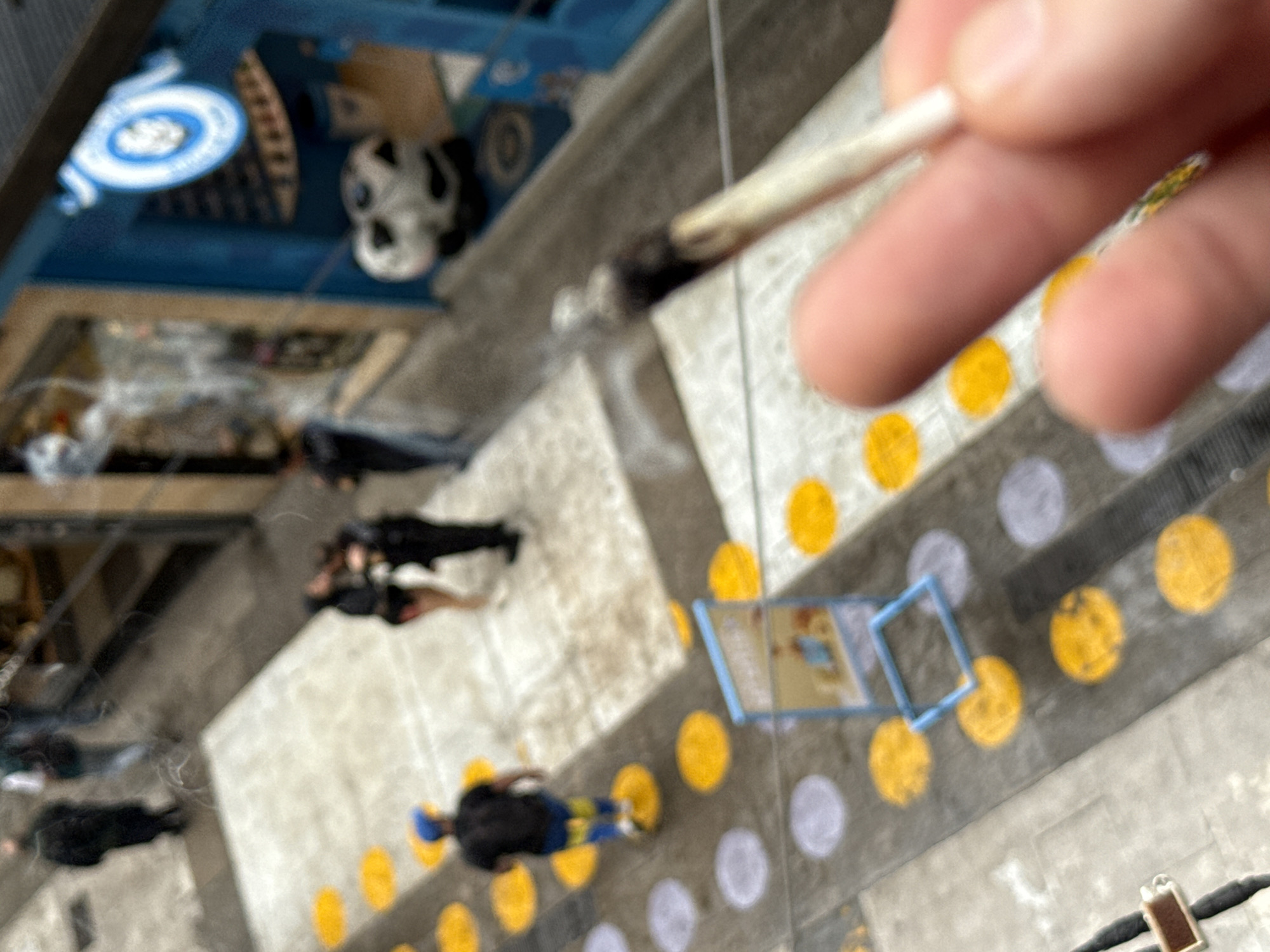 An aerial, high-angle, and generally blurry photograph captures a scene from above, overlooking a public urban space. In the immediate foreground, a person's hand is visible on the right, holding a lit, rolled item, resembling a cigarette or a joint, from which a wisp of smoke rises. The hand is partly out of focus.
Below, the ground is paved with light-colored tiles and features a pattern of painted yellow and light grey or white circles. Several indistinct figures of people are scattered across this paved area, appearing to be walking or standing casually. One person closer to the bottom left is wearing dark clothing with what looks like blue and yellow patterned pants and a blue cap.
On the ground, among the circular patterns, a light blue rectangular frame is visible, possibly outlining a game board or a sign, with faint, unreadable imagery inside.
In the upper left quadrant of the image, part of a blue overhead structure or building element is visible. Within this blue structure, a white and blue circular emblem, possibly resembling a target or a stylized logo, is discernible, along with a white object that appears to be a skull mask or sculpture. The entire scene is shot with a tilted perspective, contributing to the dynamic yet hazy quality of the image. The time of day appears to be daytime, with ambient light illuminating the scene. No specific textual information is clearly legible in the image due to the blur and distance.
