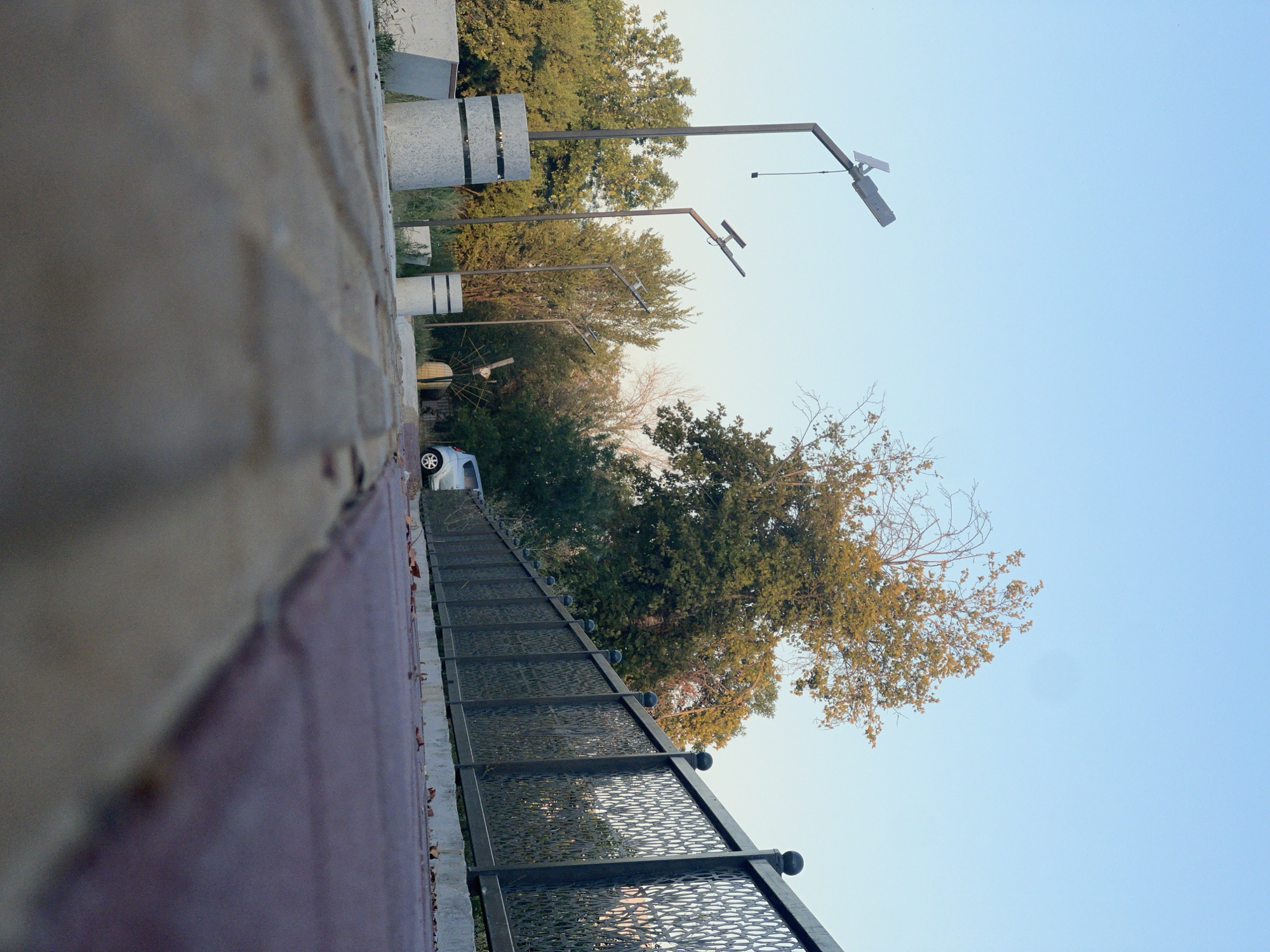 The image depicts an outdoor scene, possibly a park or promenade, with a clear blue sky overhead. In the foreground, the perspective is low, showing a pathway made of concrete and brick. To the left, there are modern-style lampposts with solar panels, and cylindrical concrete planters. A decorative metal fence with an intricate pattern runs along the middle ground, separating the pathway from a green area with trees. A silver car is parked on the right side of the scene. The environment appears to be in a city, with evidence of urban landscaping and amenities. The overall impression is one of a well-maintained public space. The scene may be located in Tashkent, Uzbekistan, given the context provided.