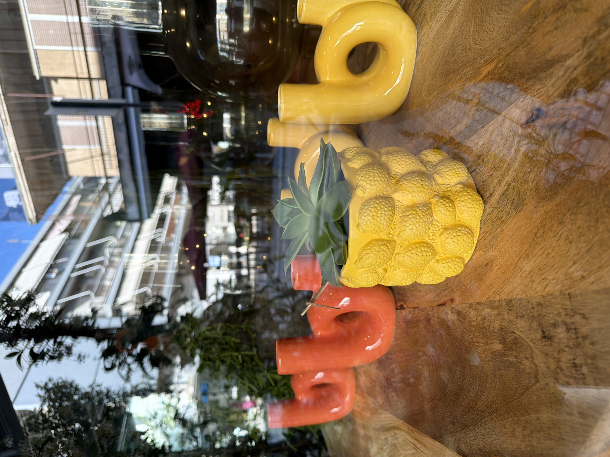 A still-life composition behind glass, likely a storefront window display in Alexandroupolis, Greece, shows three ceramic objects on a wooden surface. From top to bottom, they are a yellow number "9," a yellow pineapple-shaped container with a succulent growing out of it, and an orange "S" shaped object. The exterior is reflected in the glass, showing buildings, trees, and the sky. The color palette is dominated by yellow, orange, and shades of brown.