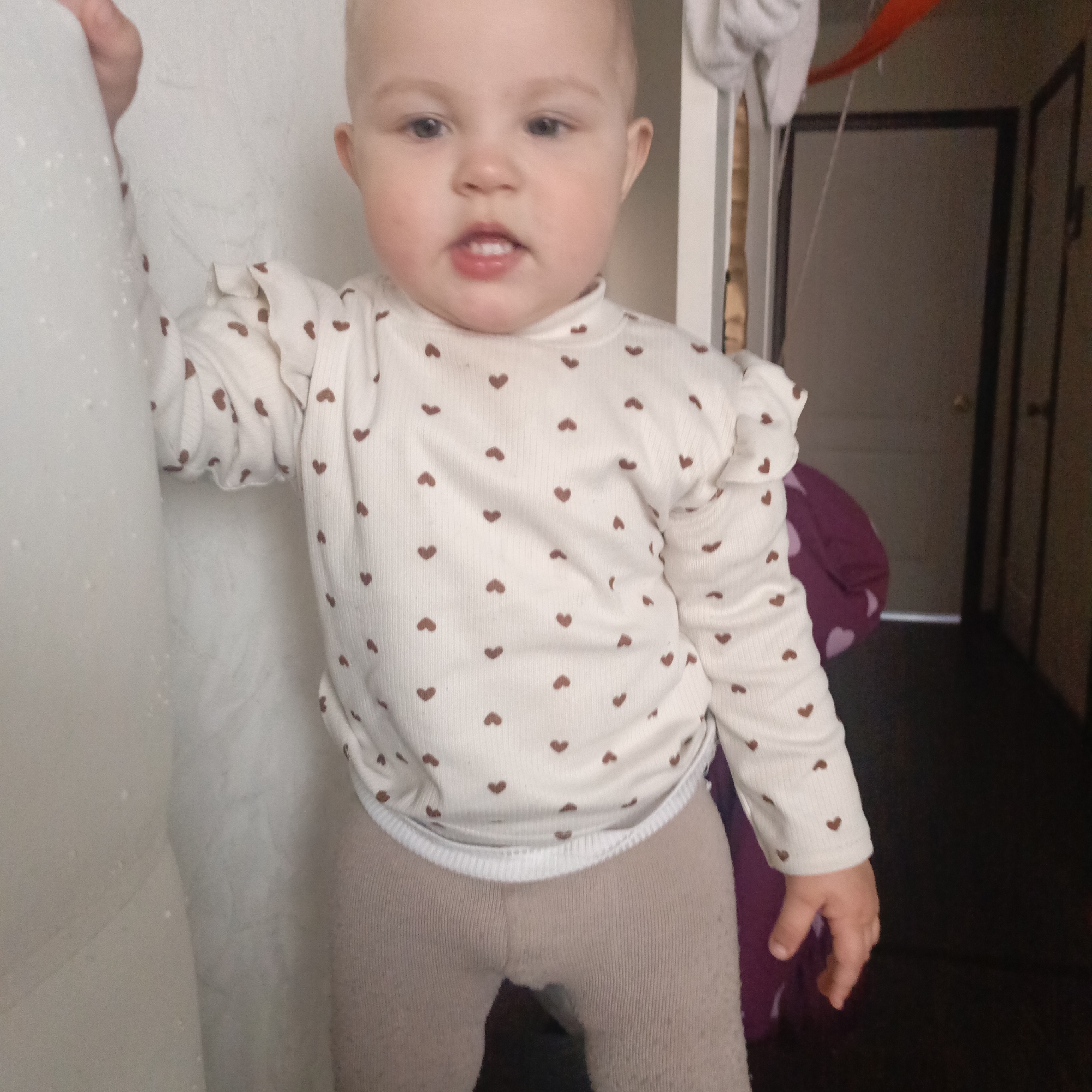 A young child, likely a baby or toddler, is captured standing indoors, looking directly at the camera. The child has fair skin and light hair, which is cropped short or sparse on top. They are wearing a cream-colored long-sleeved top adorned with numerous small, scattered brown hearts. The top features a mock turtleneck collar and ruffled accents on the shoulders. Below the top, the child wears light brown or beige ribbed leggings.
The child is positioned to the left of the frame, holding onto a white, textured surface, possibly a wall or a curtain. Their mouth is slightly open, revealing two visible bottom teeth. In the background to the right, a darker area is visible, which includes a brown door with a dark frame and a doorknob. Above and to the left of the door, some indistinct objects are hanging, including what appears to be a white cloth and a piece of orange fabric or strap. A crumpled purple or magenta object is partially visible behind the child's right side, near the floor. The overall setting suggests a casual, indoor home environment. No text is visible in the image.