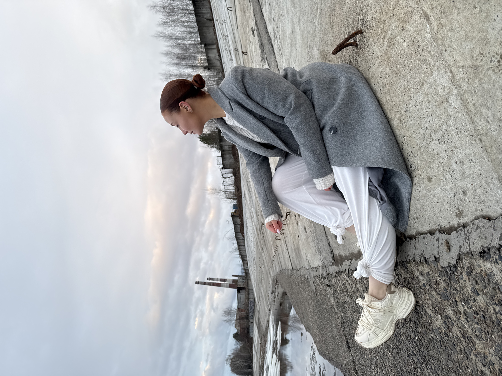 A young woman is captured in a dynamic, low-angle shot, crouching on a concrete surface in an outdoor setting in Chaikovsky, Russia. Her light brown hair is styled in a neat bun, and she has visible ear piercings. She is dressed in a warm grey overcoat layered over a white or light-colored sweater, paired with loose-fitting white pants tied at the ankles, and chunky white sneakers. Her posture suggests she is looking down or observing something intently, her gaze directed towards the right side of the frame.
The environment appears cold, likely in late autumn, winter, or early spring, evidenced by the bare trees in the background and patches of ice on a body of water. The sky is overcast, indicating a muted, diffused daylight. The setting features rugged concrete structures, possibly a riverbank or abandoned industrial area, with visible rebar and metal elements. In the mid-ground, two tall industrial chimneys or smokestacks rise, further emphasizing the industrial context of the scene. The concrete ground she is on has a coarse, textured appearance. No discernible text is visible in the image.