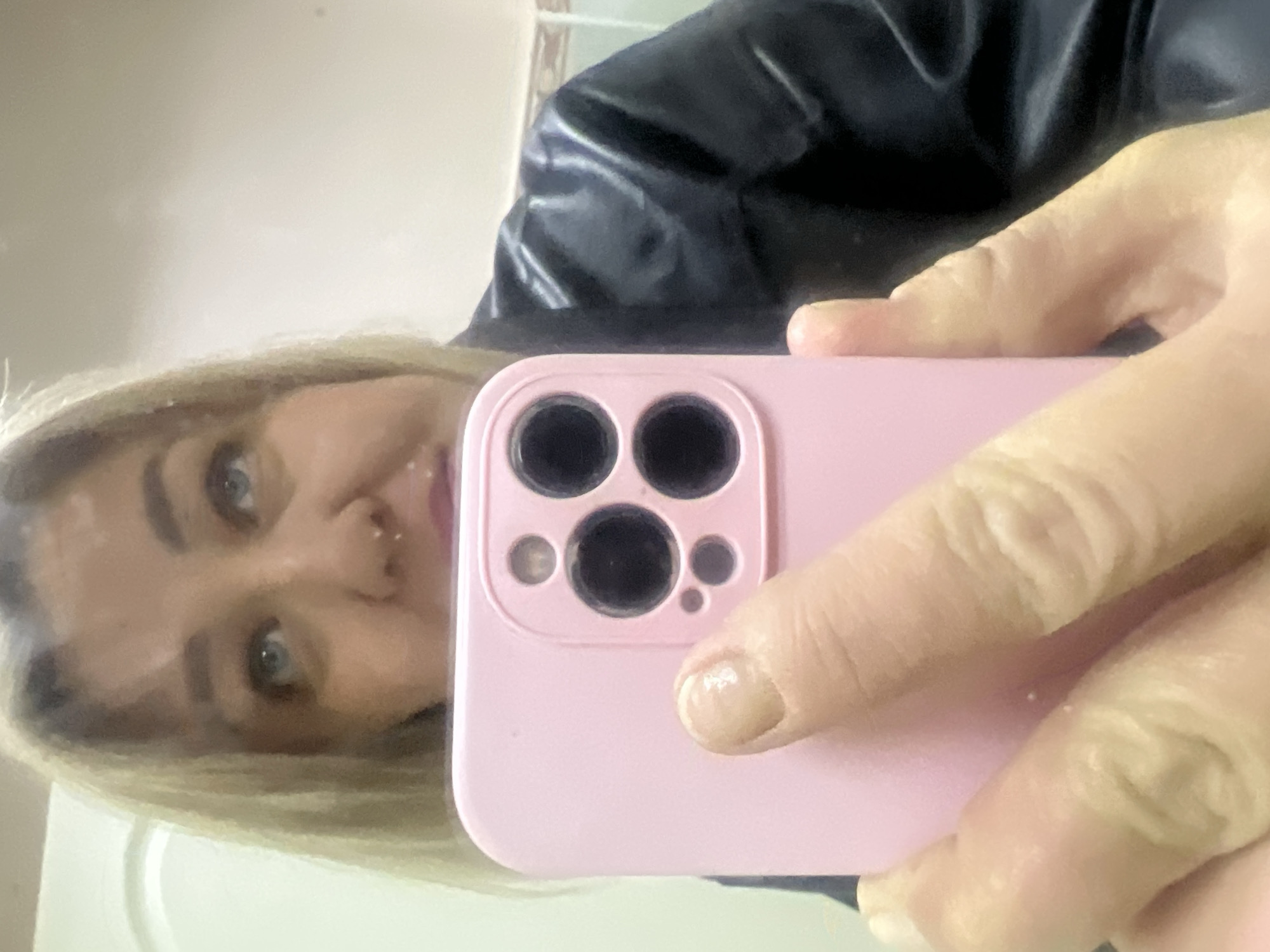 A woman with light blonde hair and blue eyes is taking a selfie with a pink smartphone. She is holding the phone with her fingers visible. A black leather jacket is partially visible in the background. The location is unknown and there is no information in the image to determine if it is in the city of Chaikovsky, Russia.