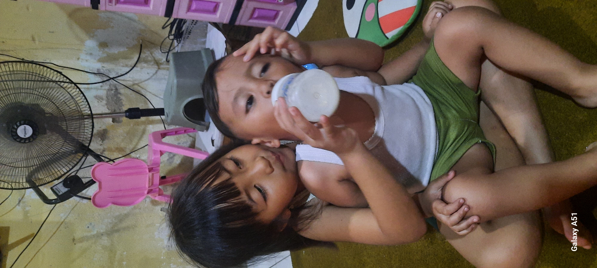 In Jatitengah, Indonesia, a young girl with dark hair and bangs tenderly holds a baby boy. The boy, wearing a white tank top and green shorts, is drinking from a white baby bottle filled with milk. The girl's left arm is around the boy's chest, and her right hand rests on his forehead. The scene takes place indoors, with a wall in the background that appears to be painted yellow and has some water stains. A black oscillating fan is visible behind the children, and a pink plastic chair with bear-shaped ears is to the left. Further back, a gray plastic object, possibly a child's stool or toy, is present. To the right, a pink multi-tiered plastic storage unit is partially visible, along with a green and white circular mat or toy on the floor. The children are sitting on a brown carpeted floor. The bottom left corner of the image displays "Galaxy A51".
