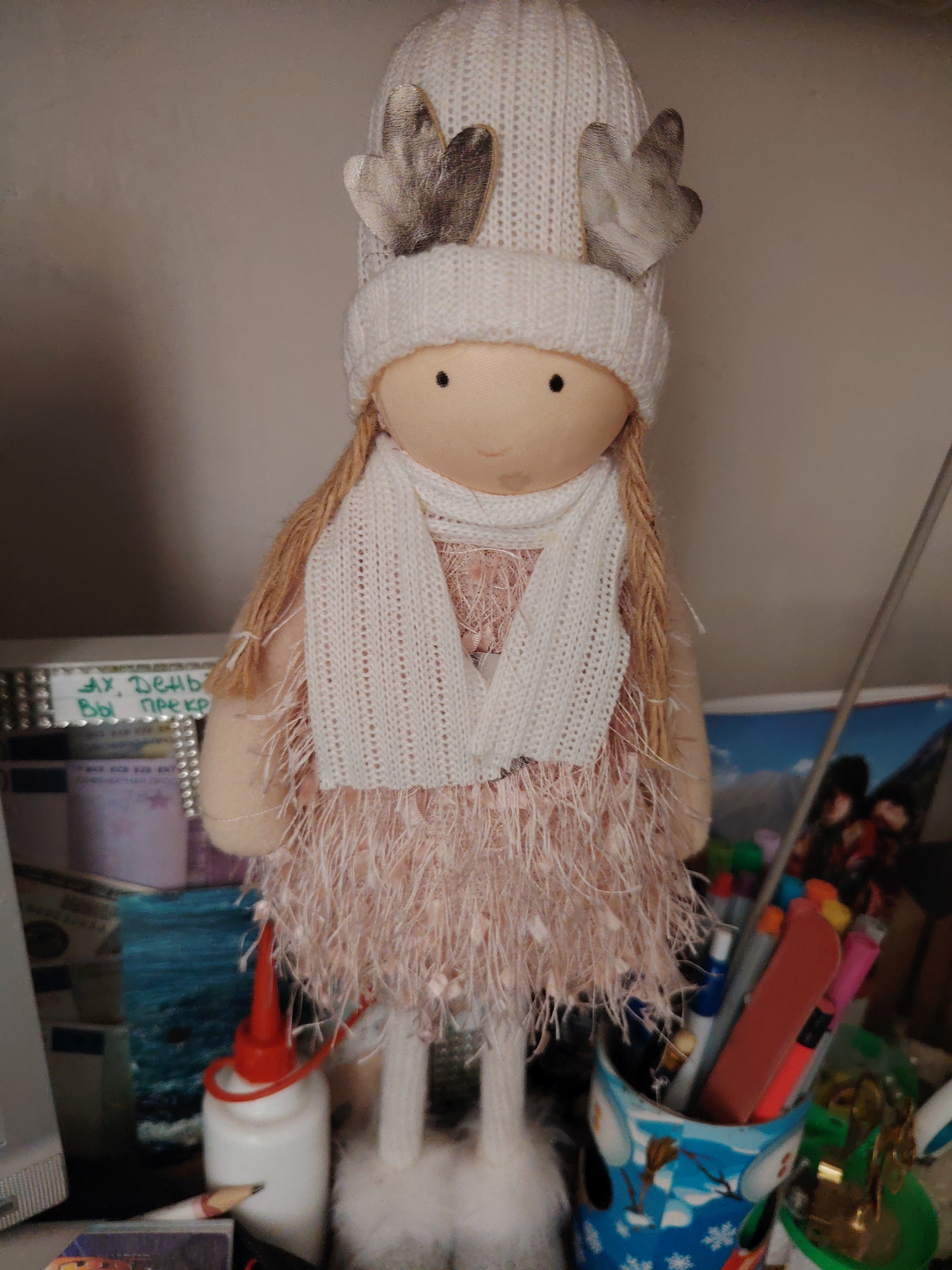 A handmade doll stands upright against a neutral background, likely a wall or shelf. The doll is dressed in a white knitted hat with what appear to be reindeer antlers made of a light-colored, textured material. It wears a white knitted scarf and a pink, feathery skirt. The doll has blonde yarn hair braided on one side and two small black eyes. A white object with a red cap, resembling a bottle of glue, is positioned to the left of the doll's feet. To the right, a collection of colorful pens and markers are stored in a blue and white patterned cup, possibly decorated with a snowman. The overall scene suggests a craft or decorative display. The location is unknown.